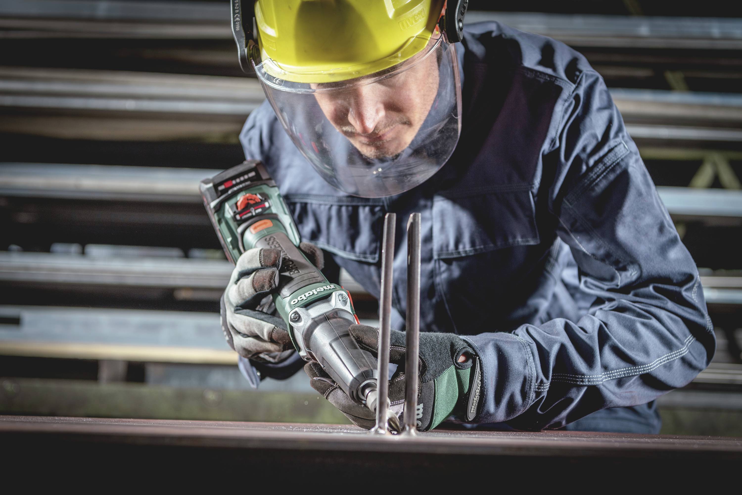 Eine Person trägt Schutzkleidung und verwendet ein elektrisches Werkzeug zum Schneiden von Metallstäben in einer Werkstattumgebung.