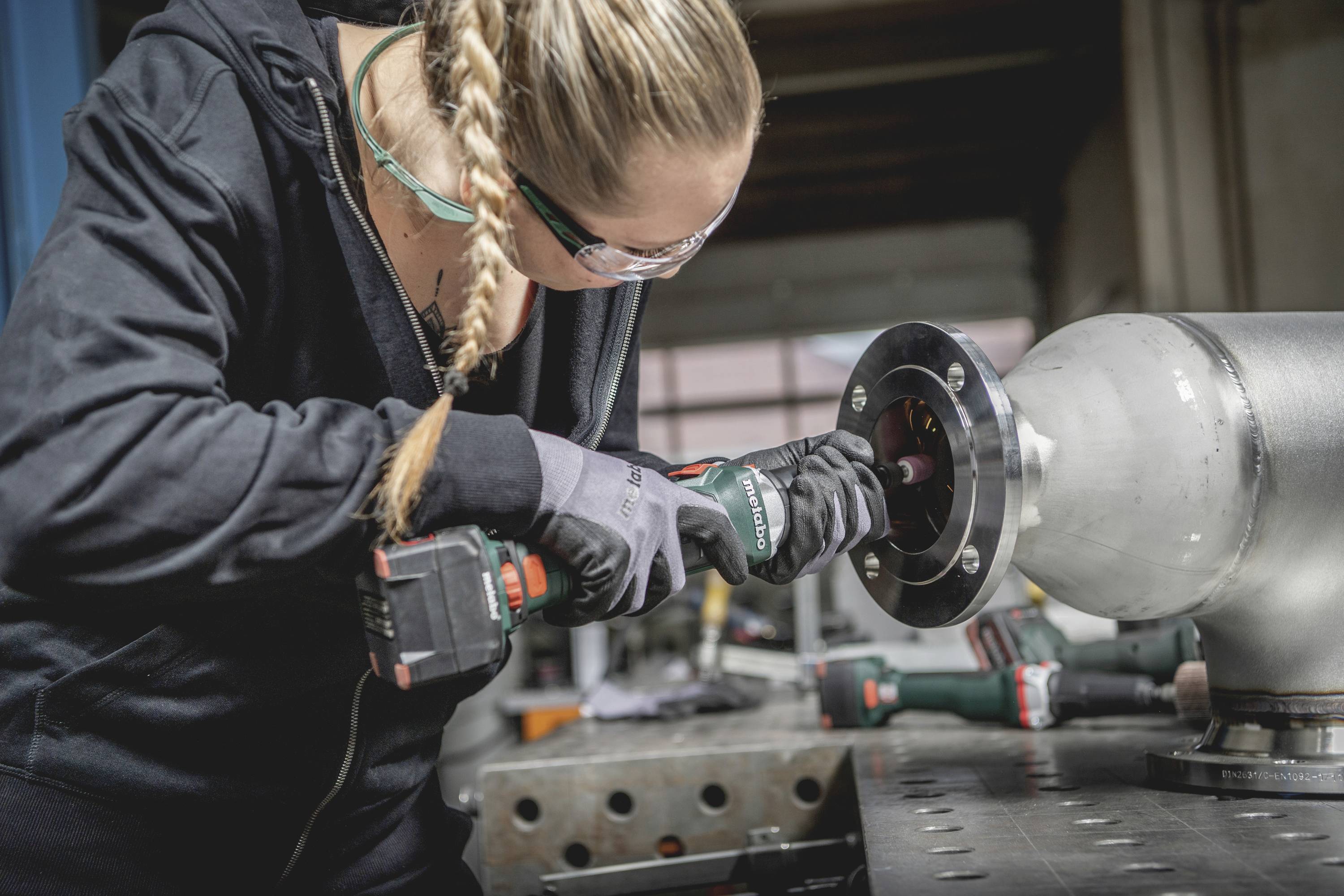 Eine Person mit Schutzbrille und Handschuhen bohrt in ein Metallstück auf einem Arbeitstisch, konzentriert bei der Arbeit.