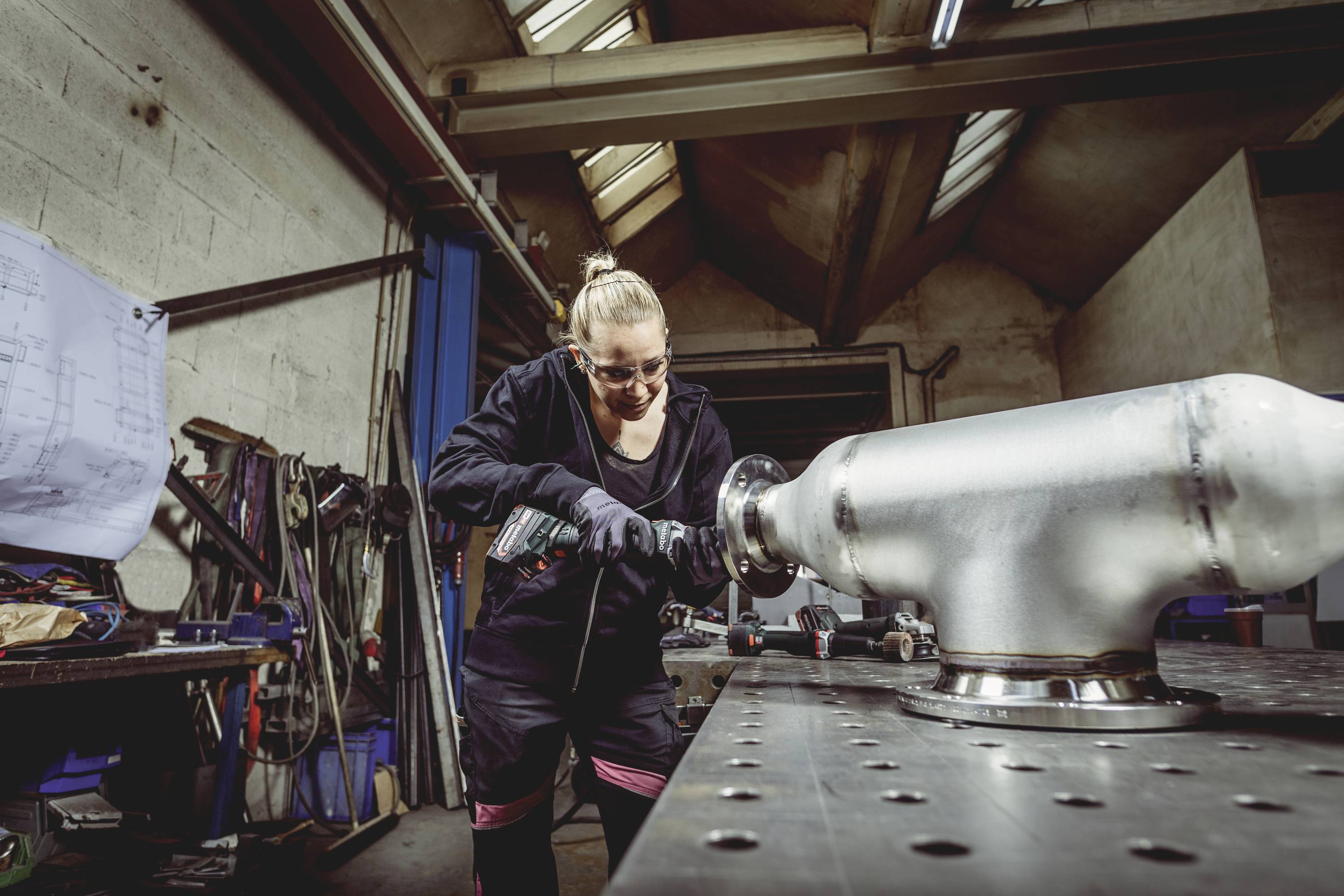 Eine Person in Arbeitskleidung bearbeitet ein großes Metallstück in einer Werkstatt mit verschiedenen Werkzeugen im Hintergrund.