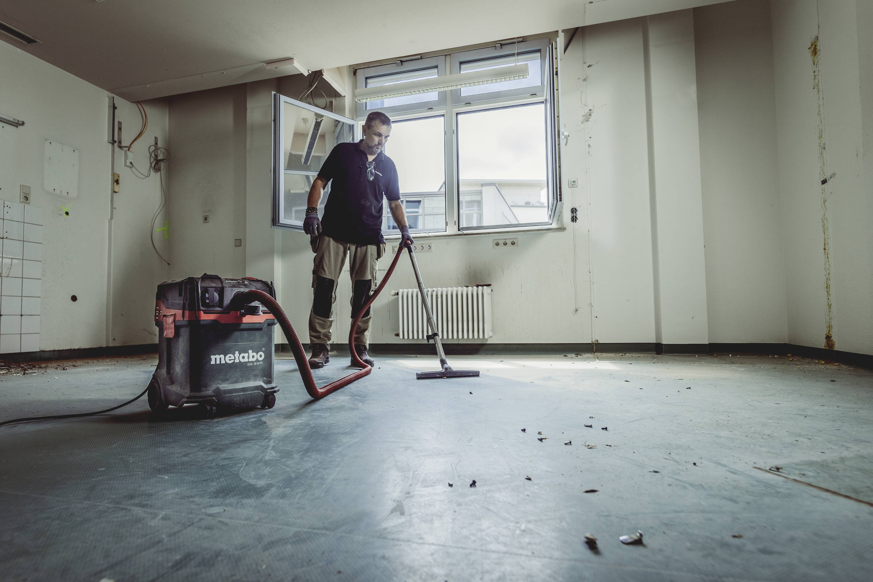 Eine Person reinigt mit einem Staubsauger einen leeren Raum mit kahlen Wänden und Fenstern im Hintergrund.
