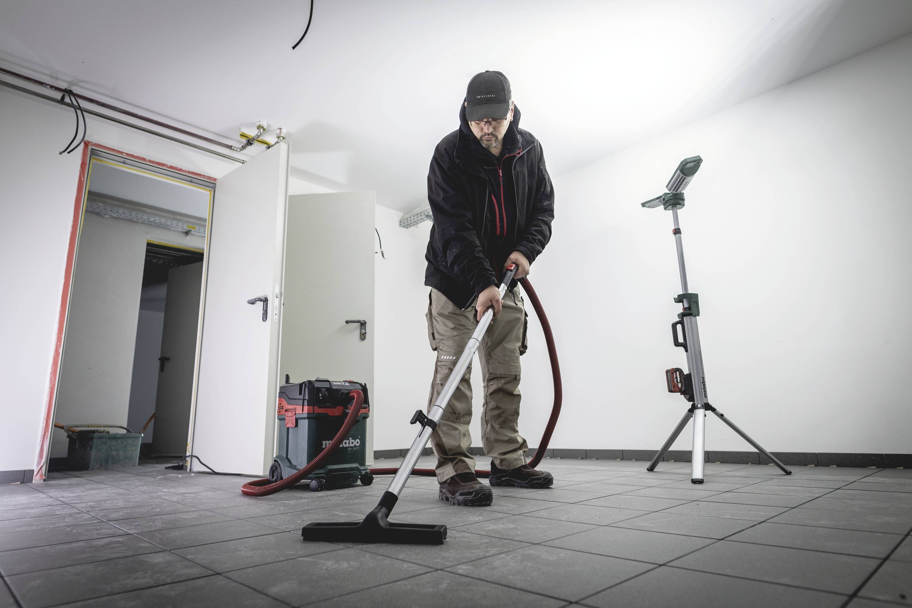 Eine Person saugt den Boden in einem leeren Raum mit einem Industriestaubsauger. Im Hintergrund ist eine offene Tür zu sehen.