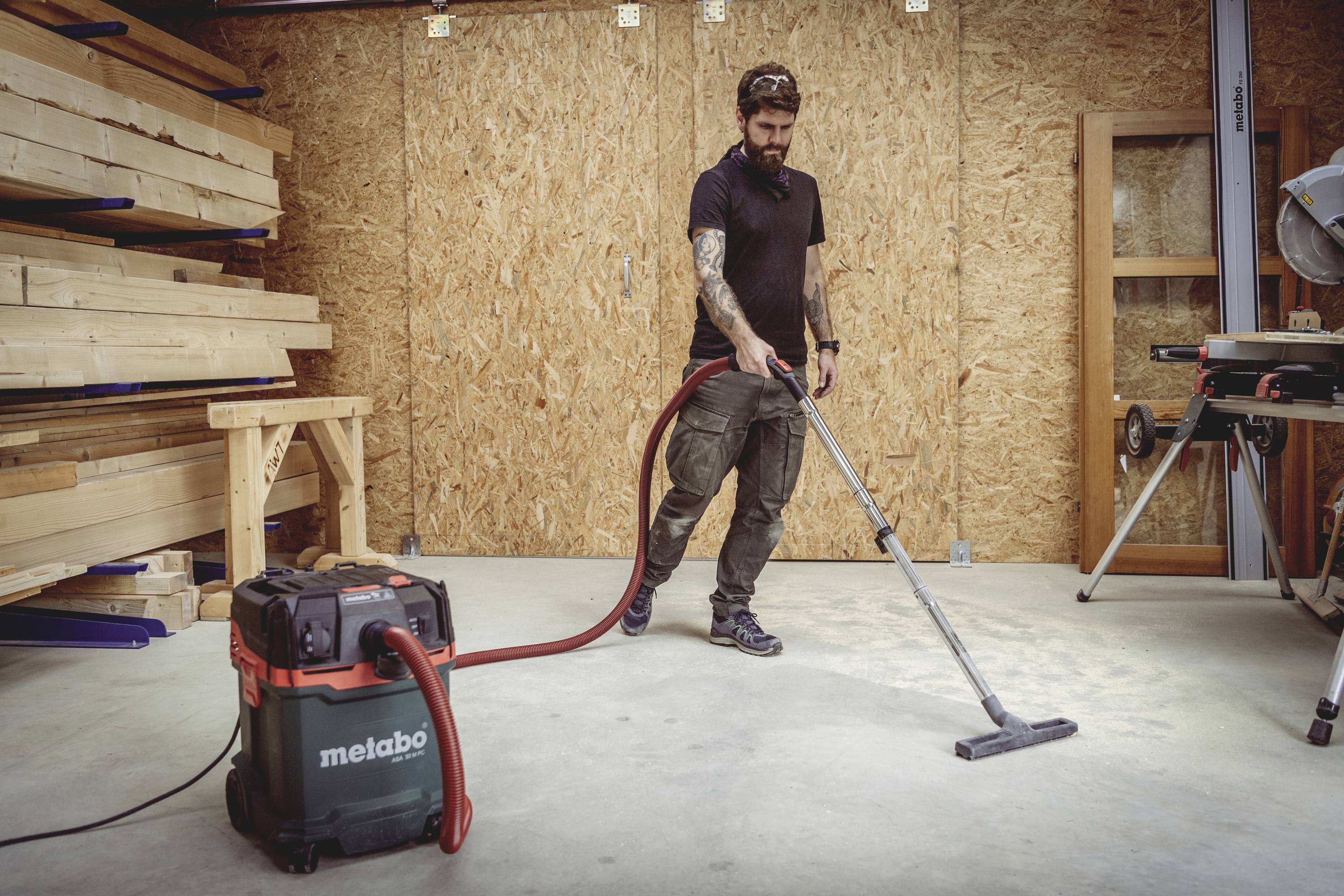 Ein Mann reinigt mit einem Industriestaubsauger den Boden in einer Holzwerkstatt. Im Hintergrund sind Holzbretter zu sehen.