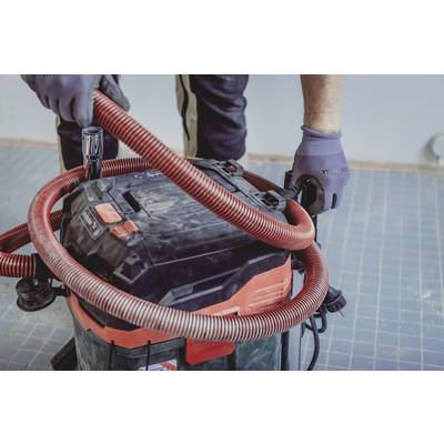 Eine Person schließt einen roten Schlauch an einen Industriestaubsauger an. Die Person trägt graue Handschuhe und arbeitet auf einem gefliesten Boden.