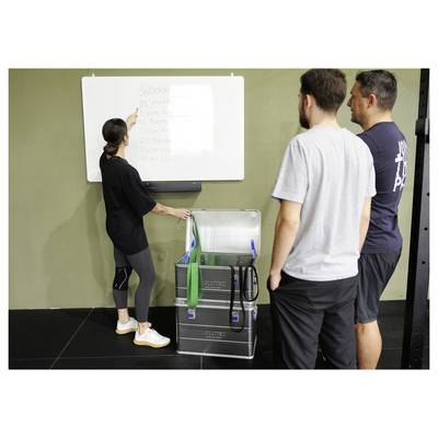 Eine Frau zeigt auf eine Liste auf einem Whiteboard, während drei Männer in der Nähe stehen, neben offenen Ausrüstungskisten, in einer Fitnessstudio-Umgebung.
