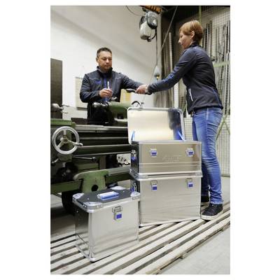 Ein Mann und eine Frau stehen in einer Werkstatt neben einer Metallmaschine und schauen sich Dokumente an. Drei silberne Lagerkisten sind in der Nähe gestapelt.