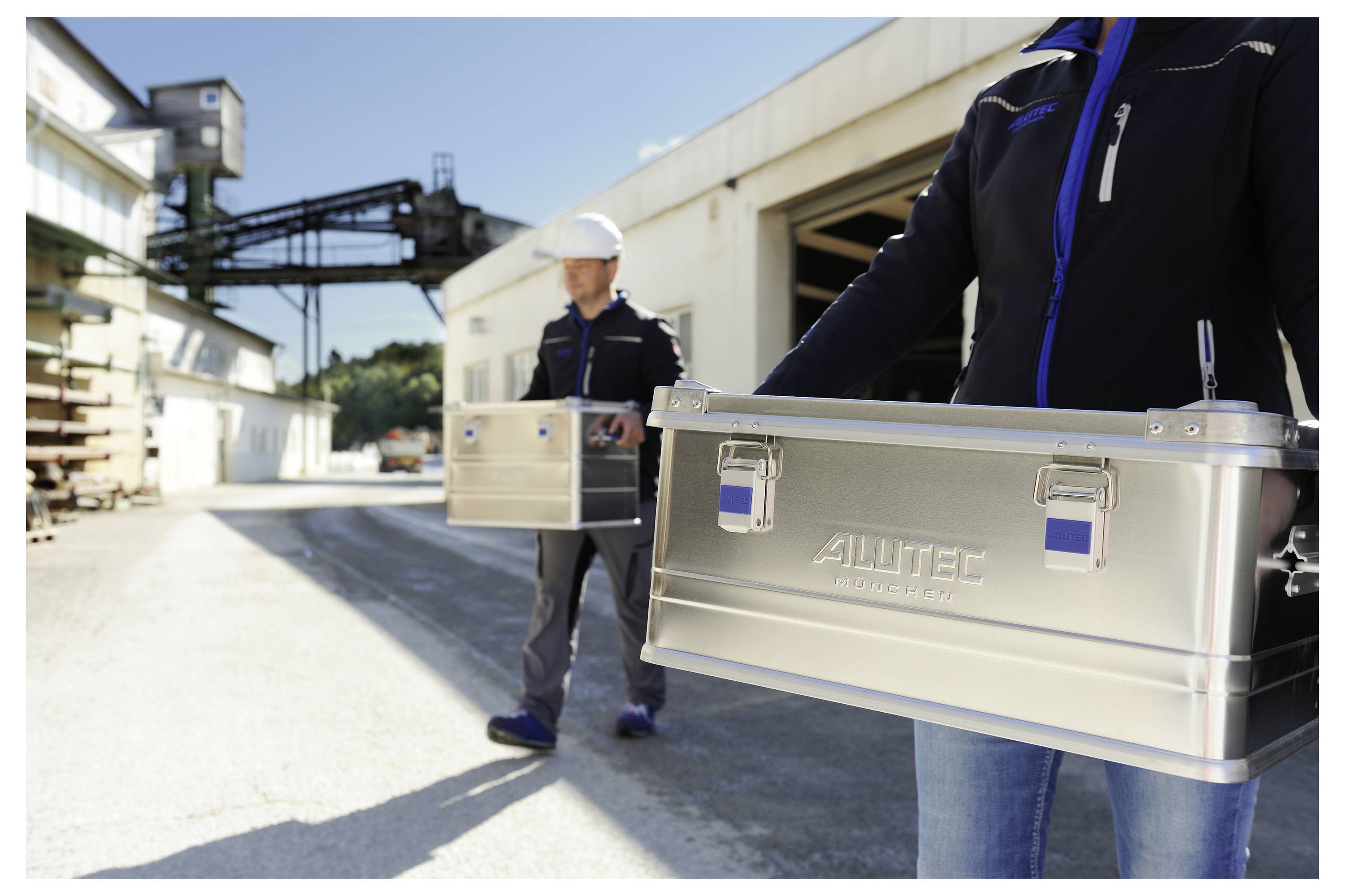 Zwei Personen tragen metallische Werkzeugkästen mit der Aufschrift "ALUTEC" im Freien auf einem Industriegelände, mit Gebäuden und Maschinen im Hintergrund.