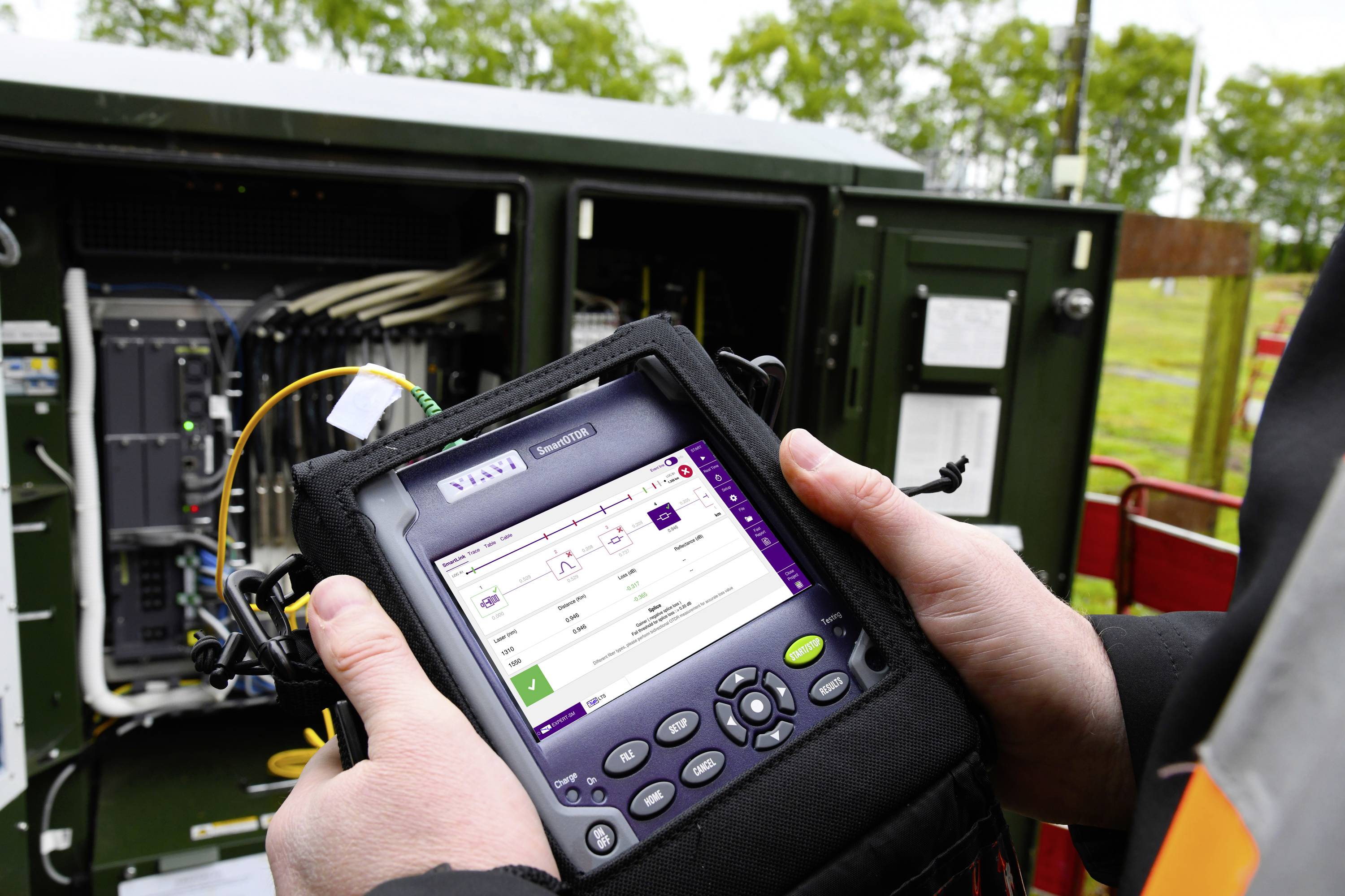Eine Person hält ein Messgerät vor einem offenen Schaltschrank im Freien. Das Gerät zeigt technische Daten an. Im Hintergrund sind Bäume sichtbar.