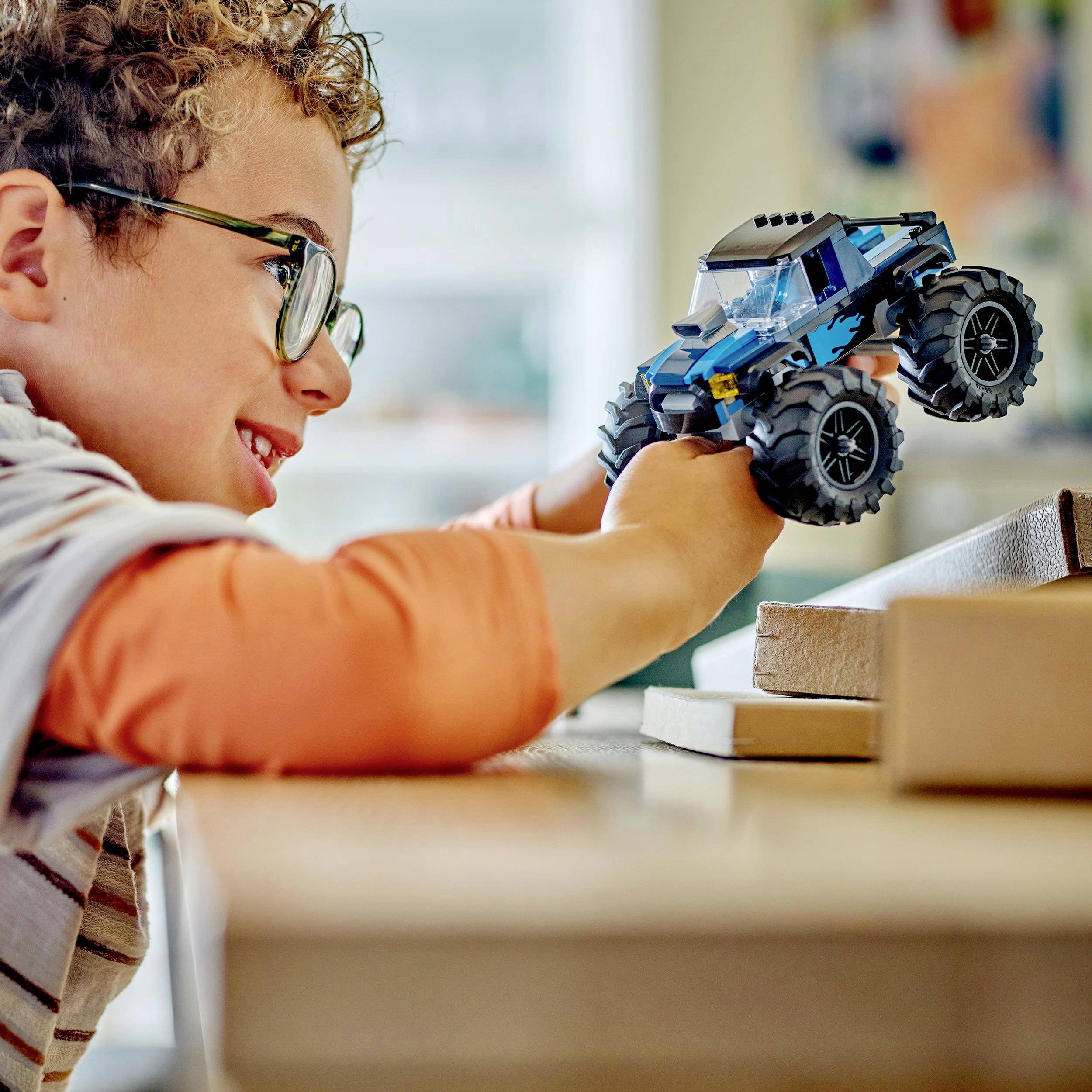 Ein Junge mit lockigem Haar und Brille spielt begeistert mit einem blauen Spielzeugauto auf einem Tisch.