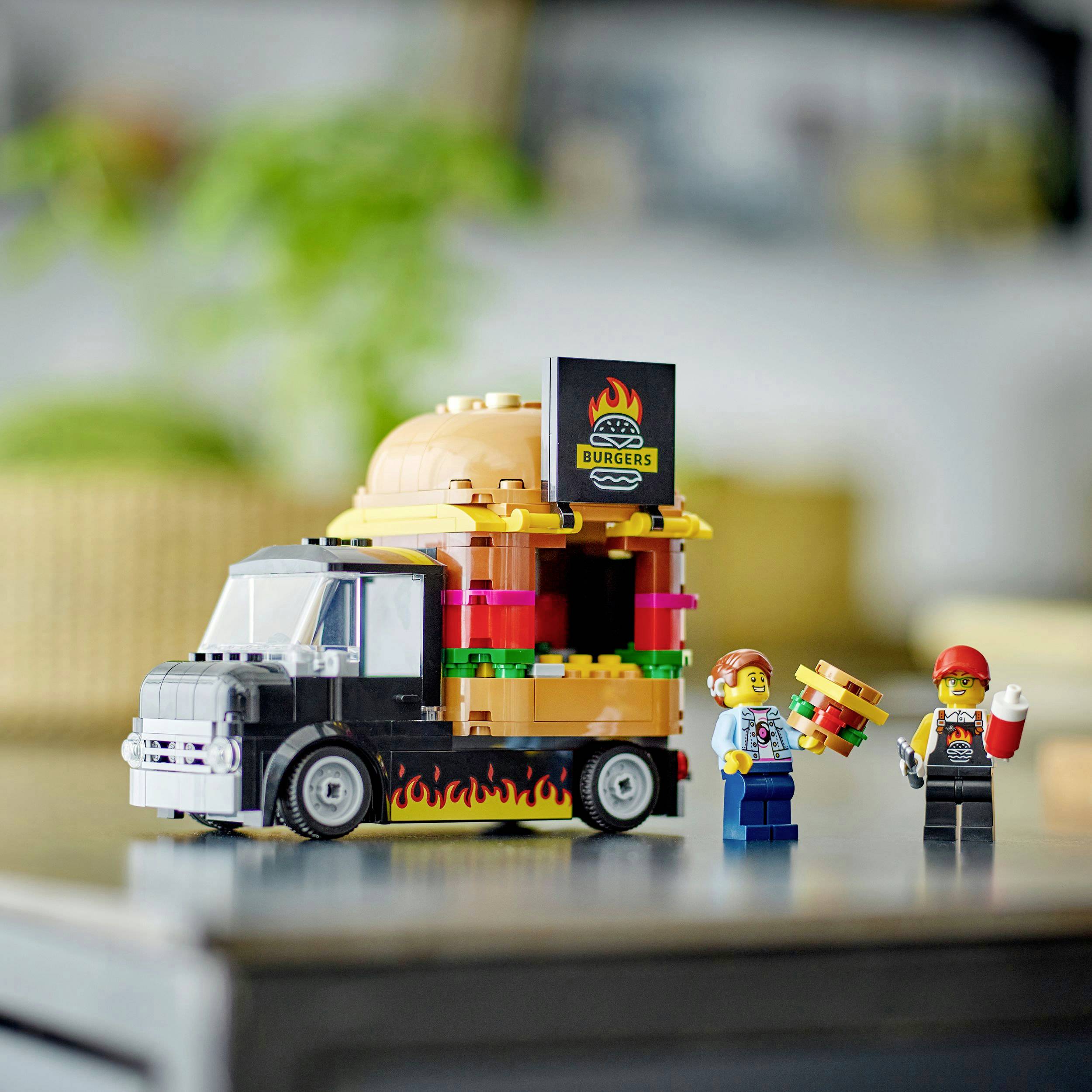 Spielzeuge Truck in Form eines Hamburgers; zwei Figuren stehen daneben mit einem winzigen Hamburger in der Hand. Bunte, lebendige Szene.