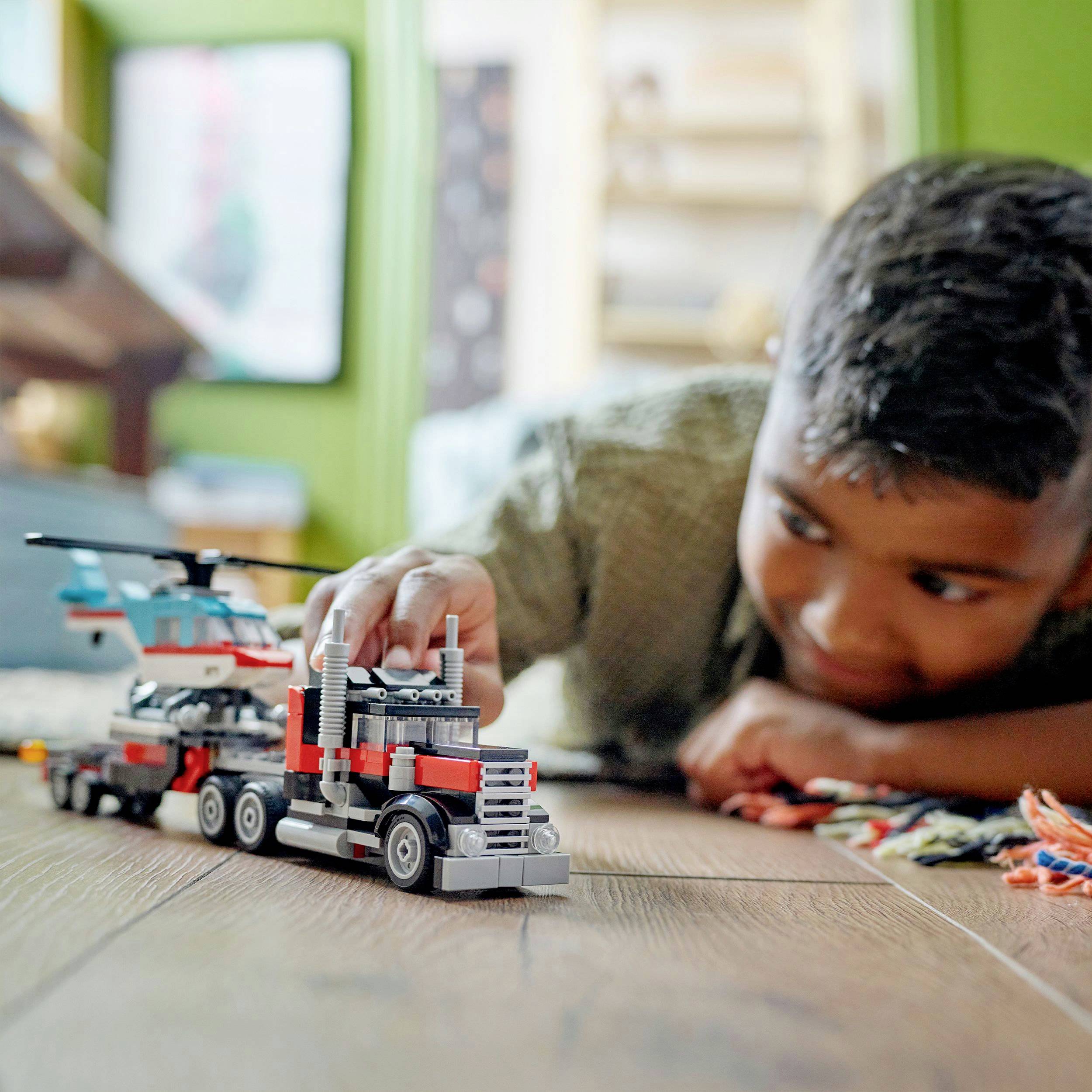Kind spielt mit Spielzeug-LKW und Hubschrauber auf dem Boden. Im Hintergrund unscharfe Möbel und Dekorationen.