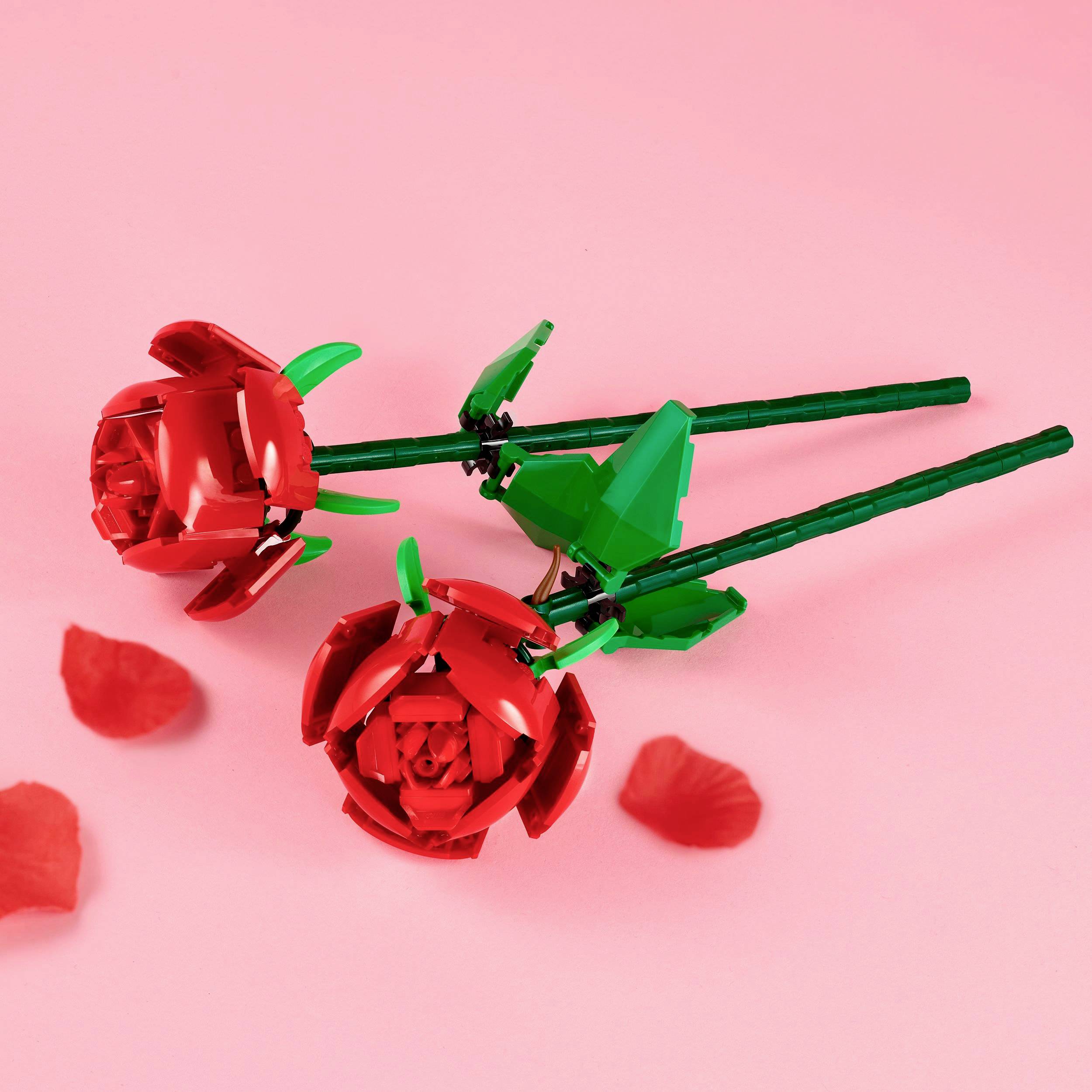 Zwei rote Rosen aus LEGO-Steinen auf rosa Hintergrund, mit grünen Stielen und Blättern. Lose Blütenblätter liegen daneben.