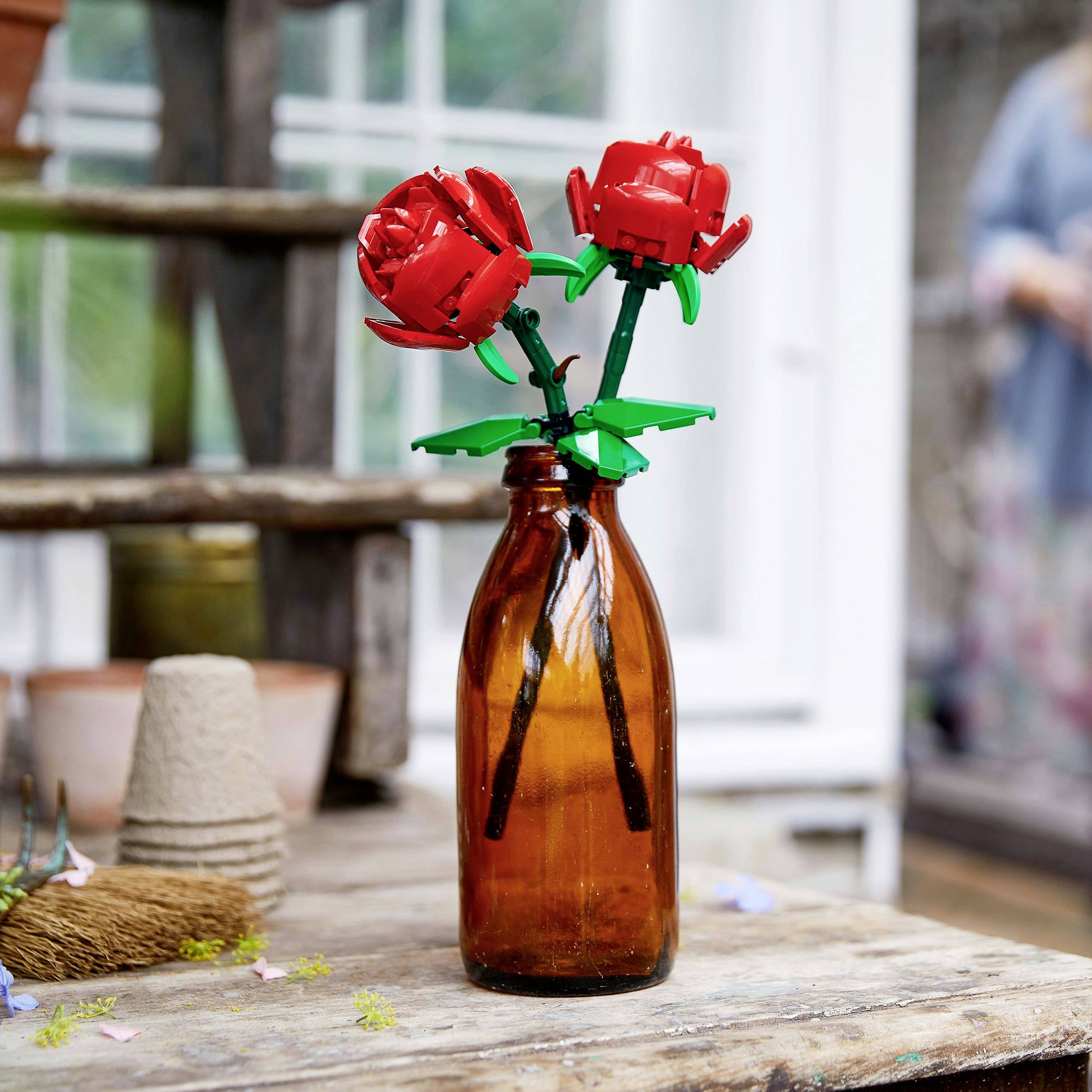 Zwei rote LEGO-Rosen in einer braunen Glasvase stehen auf einem Holztisch, im Hintergrund unscharfe Garten- und Hausmotive.