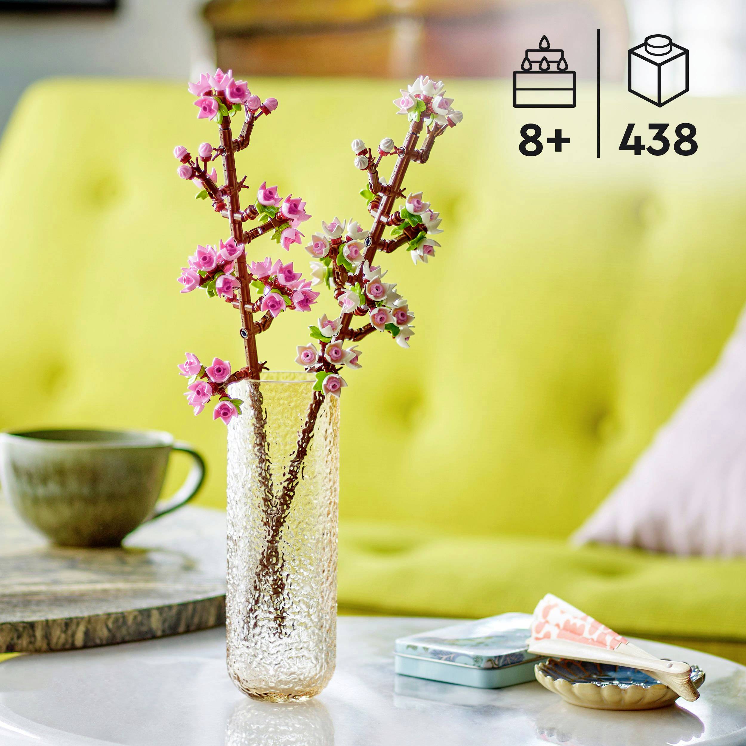 Ein dekoratives rosa Blumenarrangement in einer strukturierten Vase steht auf einem Tisch mit Teetasse und Schachtelspiel. Symbole '8+' und '438' sind sichtbar.