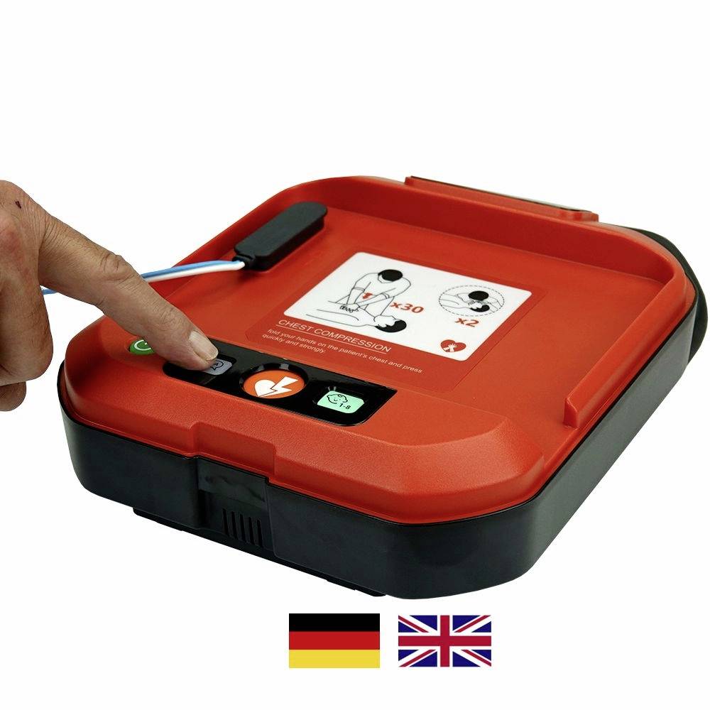 Ein Defibrillator mit einer Anleitung auf der Oberseite. Ein Finger zeigt auf eine Taste zum Starten des Geräts. Darunter Flaggen von Deutschland und Großbritannien. 