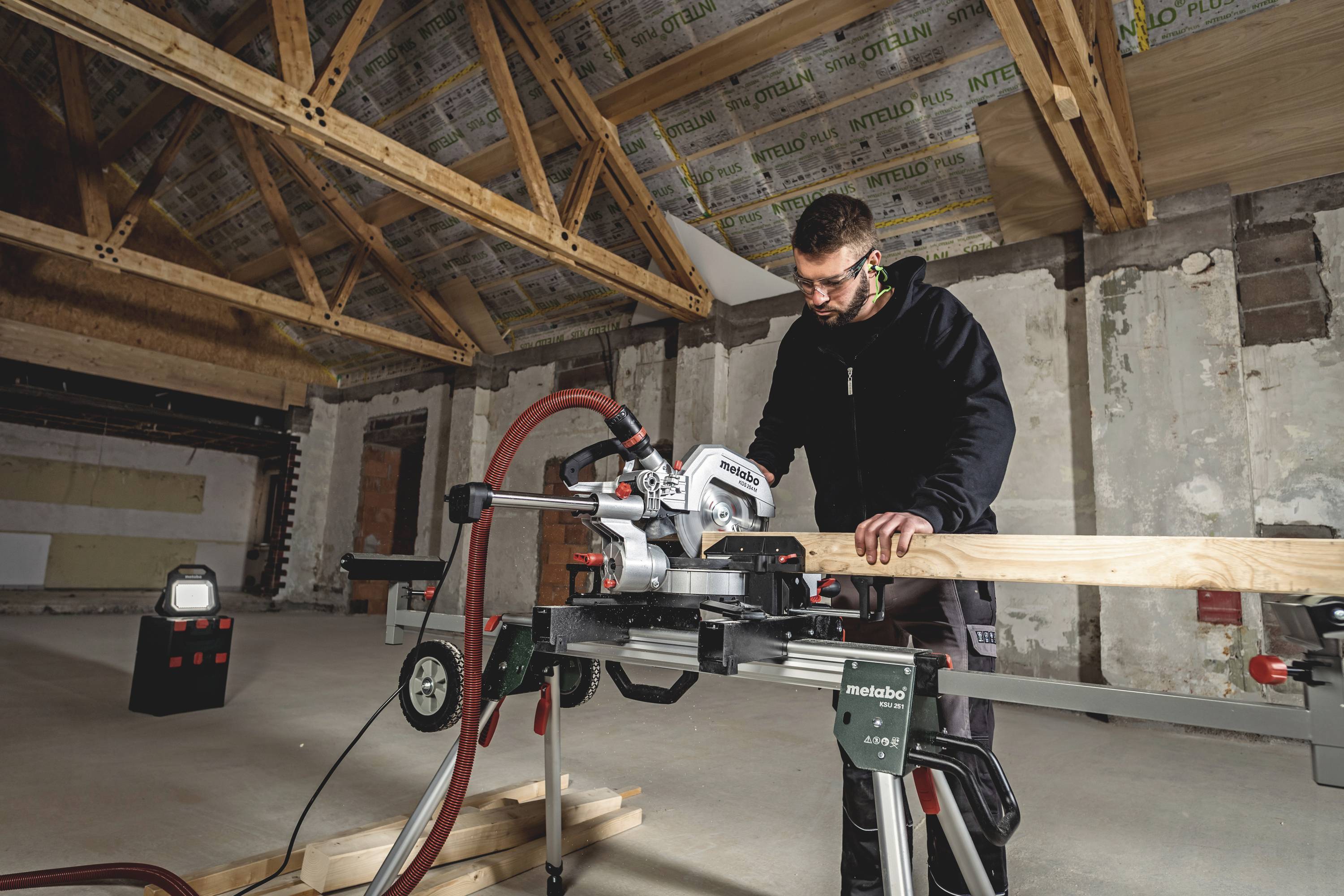 Ein Mann sägt mit einer elektrischen Säge ein Holzbrett in einem halbfertigen Raum mit sichtbaren Holzbalken und unvollständigen Wänden.