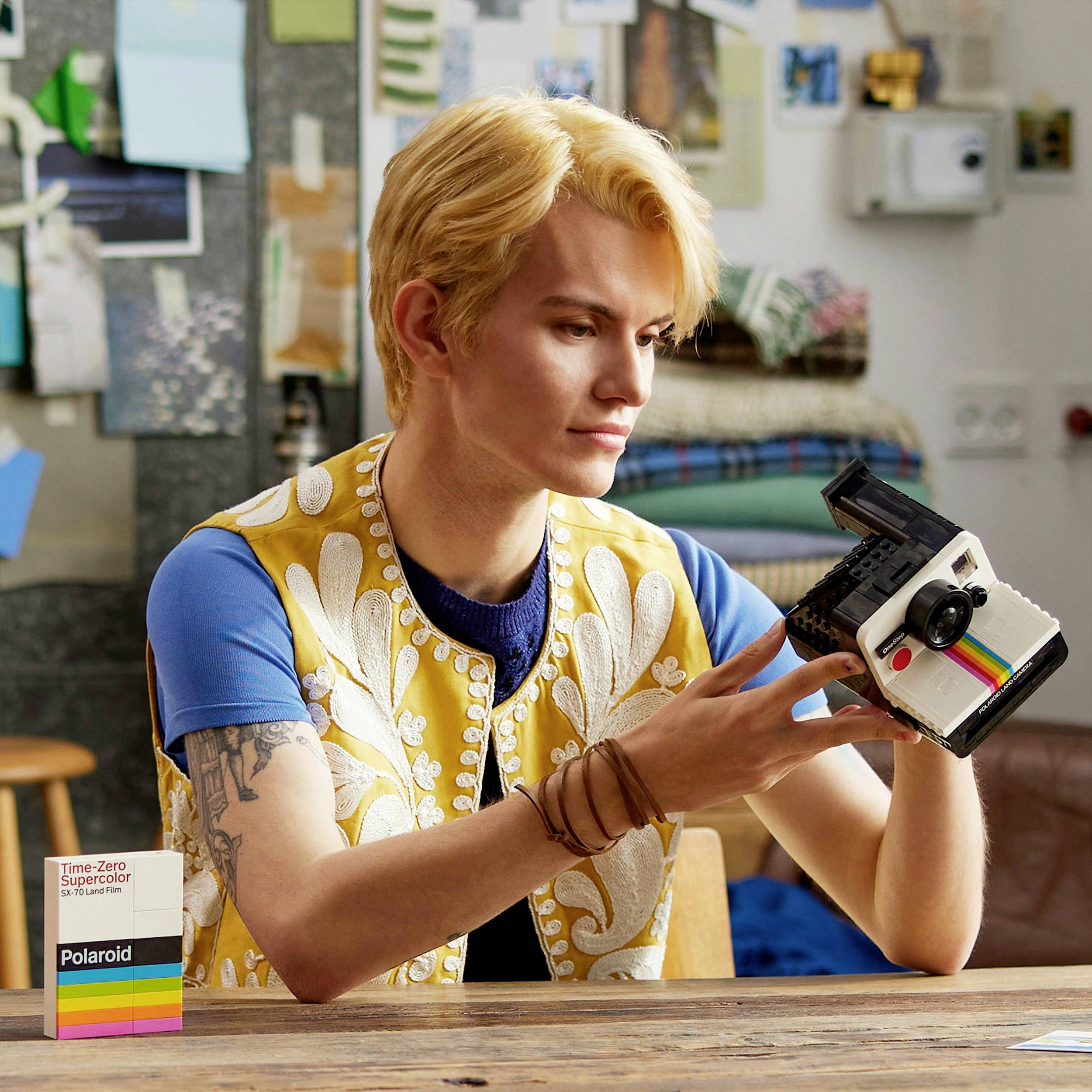 Ein Mann hält eine Polaroid-Kamera in der Hand, während er nachdenklich schaut. Im Hintergrund sind Tafel und Bilder an der Wand.