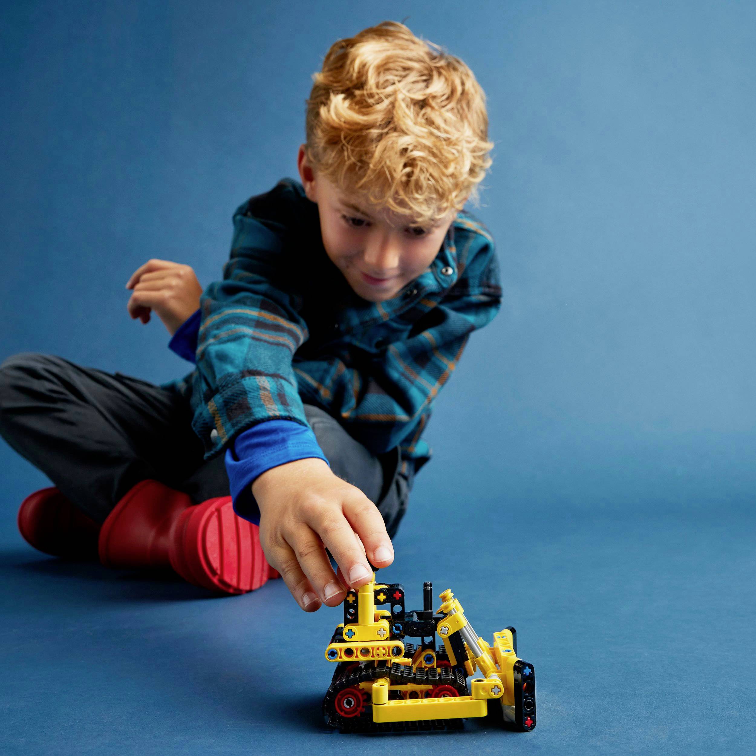 Ein Kind spielt konzentriert mit einem gelben Spielzeugbagger auf einem blauen Boden. Es trägt ein kariertes Hemd.