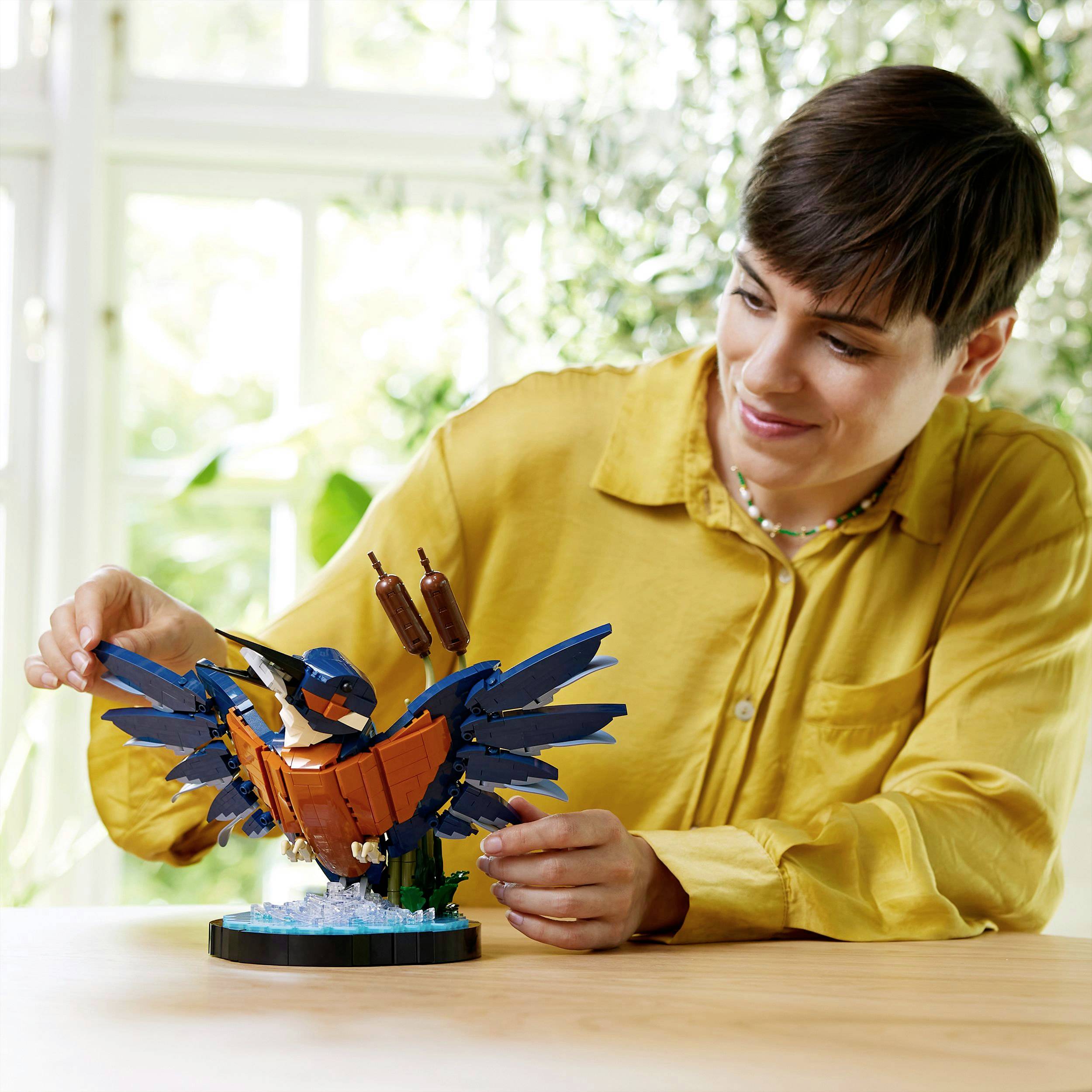 Eine lächelnde Person sitzt an einem Tisch und betrachtet ein kunstvoll gebautes LEGO-Modell eines Vogels mit blau-orangefarbenem Gefieder.