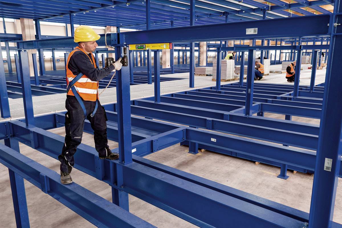 Arbeiter mit Schutzhelm und Warnweste montiert Metallregale in Lagerhalle. Nutzung einer Wasserwaage, um Präzision zu sichern. Im Hintergrund zwei weitere Arbeiter.