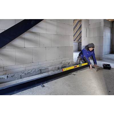 Eine Person in Arbeitskleidung nivelliert mit einer Wasserwaage einen Stahlträger in einem Rohbau. Wand und Balken im Hintergrund.