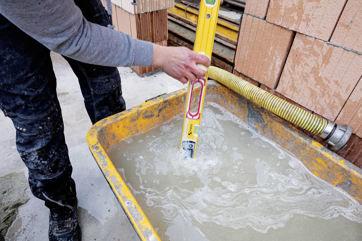 Eine Person prüft die Wasserwaage in einer gelben Betonmischschale mit Wasser. Im Hintergrund sind Ziegelsteine zu sehen.
