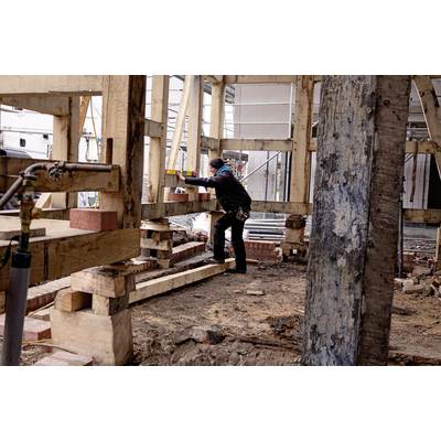 Eine Person arbeitet in einem teils errichteten Holzrahmenbau, misst mit einer Wasserwaage und trägt Schutzkleidung.