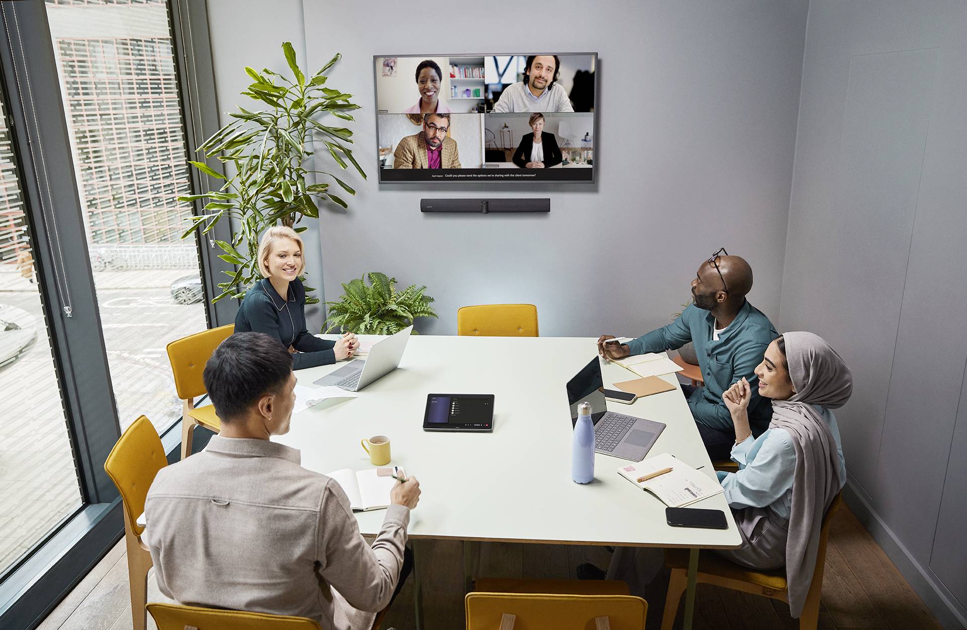 Eine Gruppe von fünf Personen sitzt in einem Konferenzraum und nimmt an einer virtuellen Besprechung auf einem Bildschirm an der Wand teil.