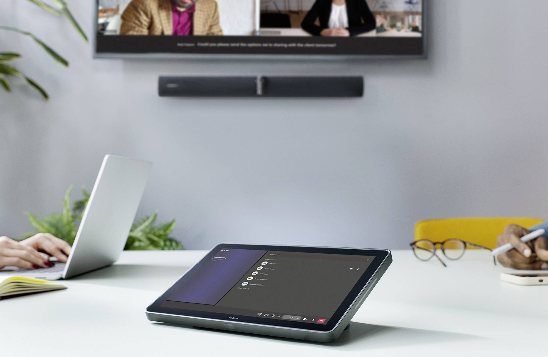 Eine Person arbeitet an einem Laptop auf einem Tisch. Im Vordergrund liegt ein Tablet mit einer Konferenzanwendung. Ein Bildschirm ist im Hintergrund.