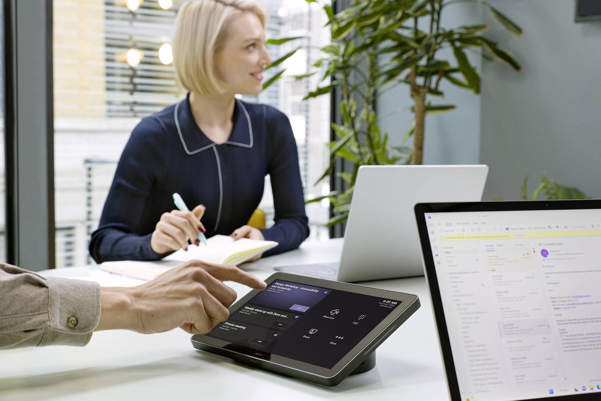 Person in einem Büro zeigt auf ein Touchscreen-Tablet auf einem Tisch, während eine andere Person am Laptop sitzt und Notizen macht.