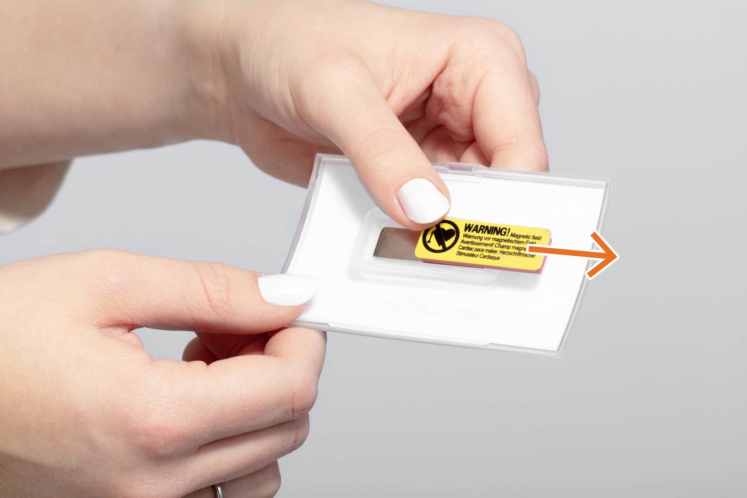 Zwei Hände halten eine Kunststoffhalterung mit einem gelben Warnaufkleber darauf. Der Aufkleber befindet sich in der Mitte der Halterung.