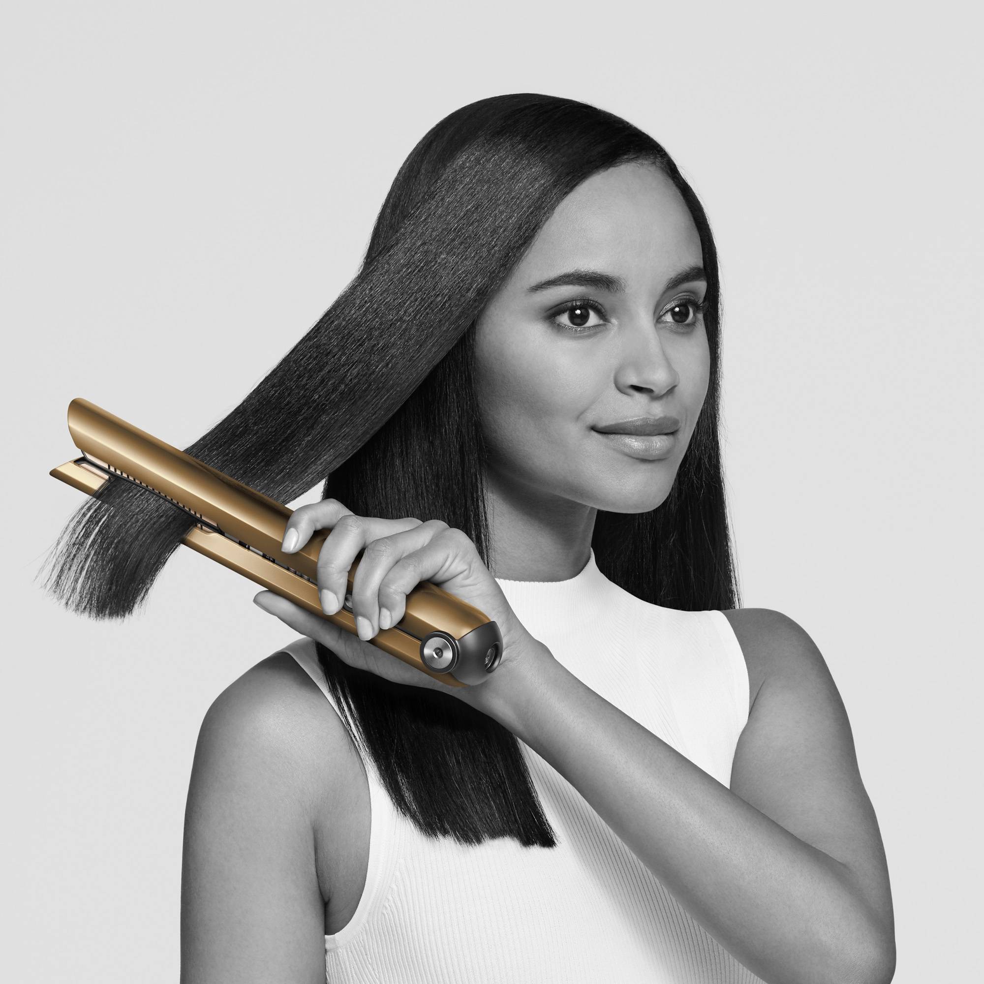 Eine Frau glättet ihr langes, dunkles Haar mit einem goldfarbenen Glätteisen. Sie trägt ein ärmelloses, weißes Oberteil.