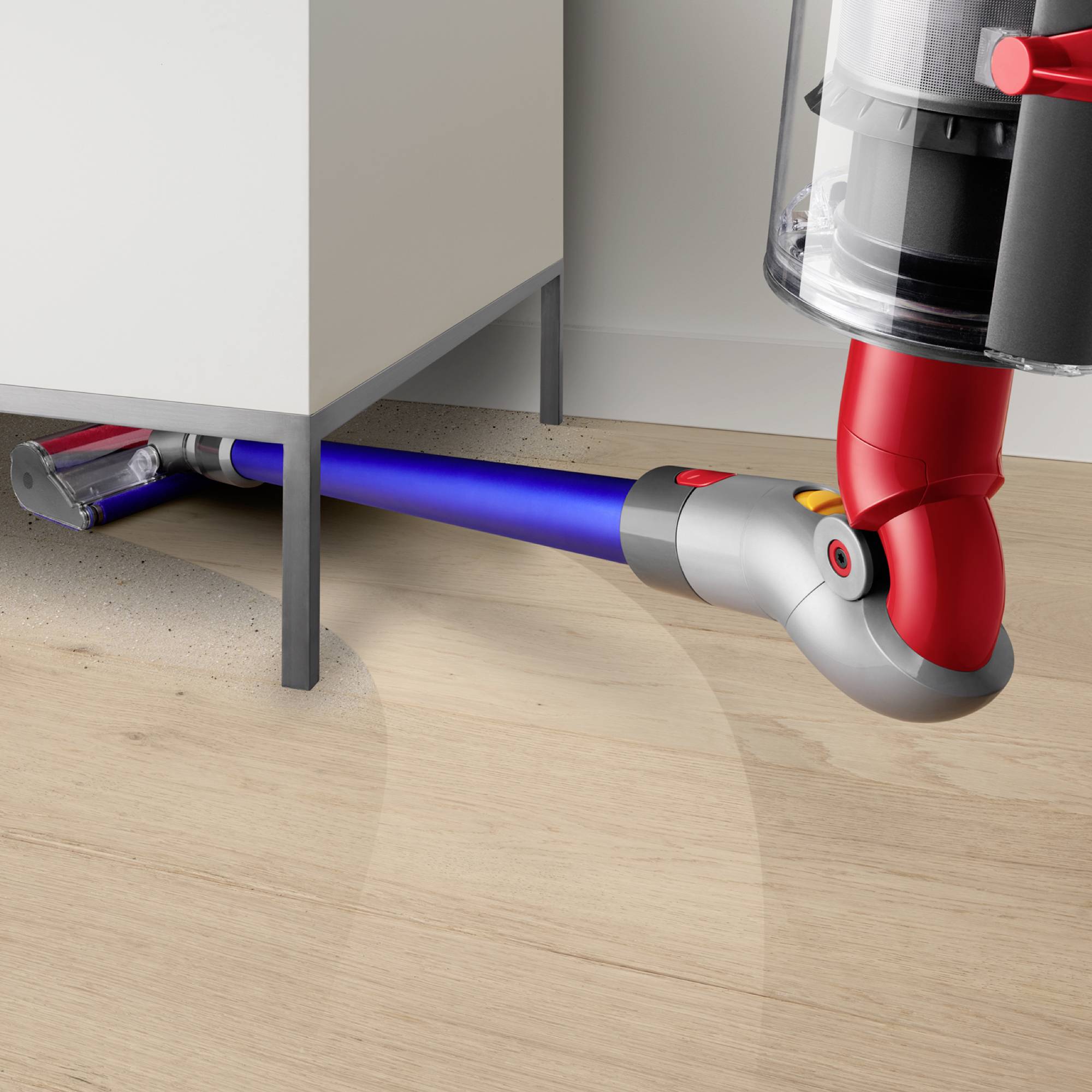 Ein roter und grauer Akkusauger saugt mit einem blauen Schlauch Schmutz unter einem weißen Schrank auf hellem Holzboden auf.