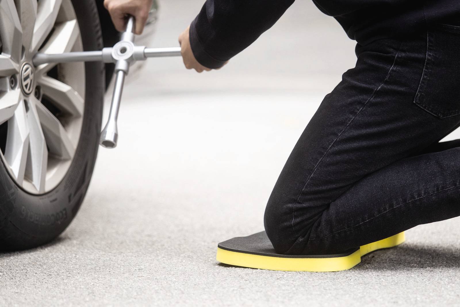 Person kniet auf Kniepolster und wechselt Autoreifen mit Reifenheber auf einer Straße.