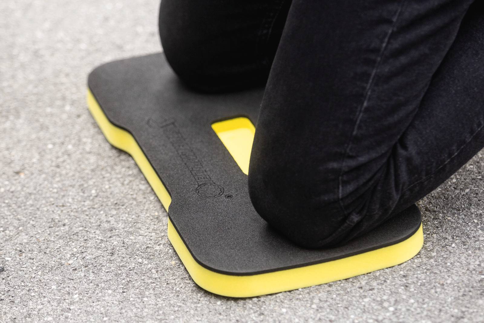 Eine Person kniet auf einem schwarzen Kniekissen mit gelbem Rand auf einem Asphaltboden.