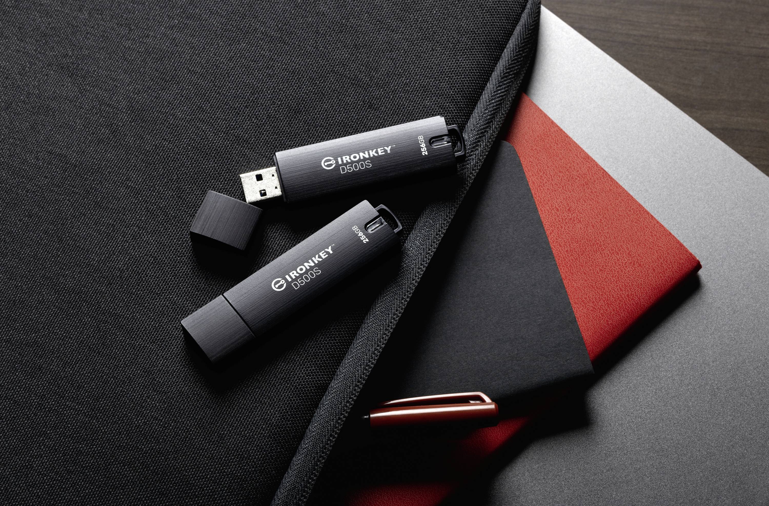 Zwei USB-Sticks mit der Aufschrift 'IronKey D300S' auf einer schwarzen Tasche neben roten und schwarzen Notizbüchern und einem Stift.