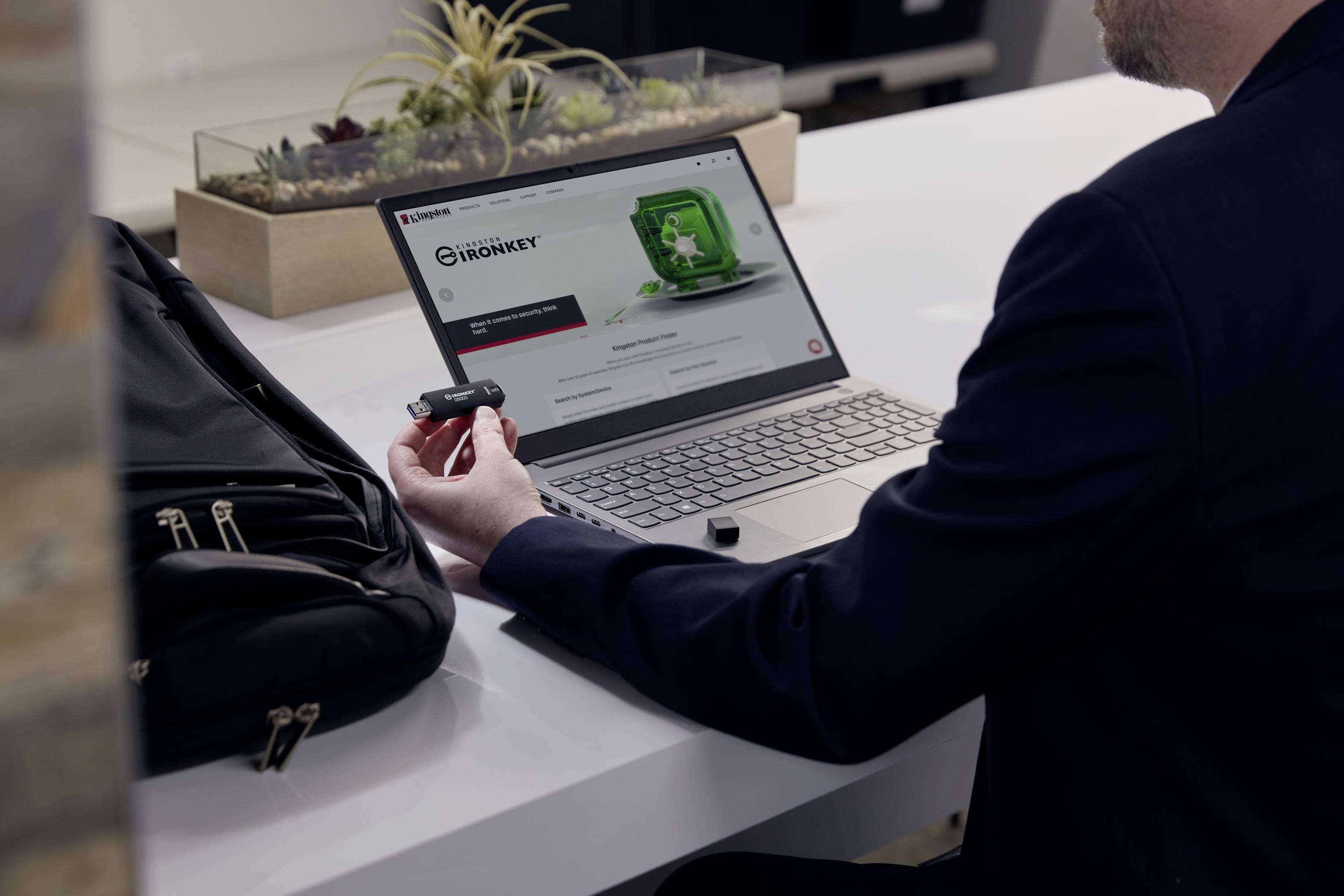 Ein Mann sitzt an einem Schreibtisch und hält einen USB-Stick, während er eine Webseite auf einem Laptop ansieht. Neben ihm liegt ein Rucksack.
