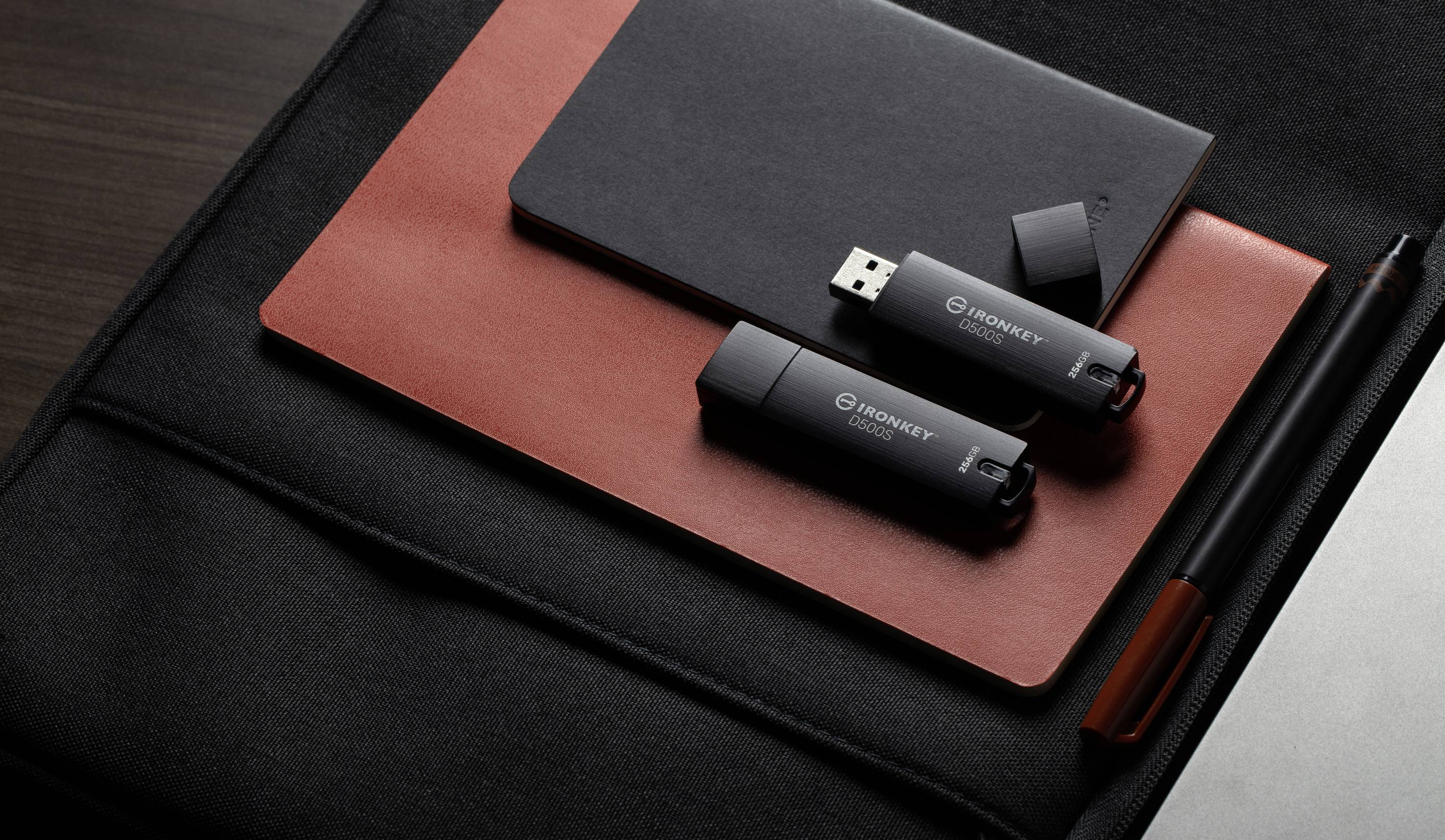 Zwei schwarze USB-Sticks liegen auf einem roten Notizbuch und einem dunklen Laptop in einer eleganten Büroumgebung.