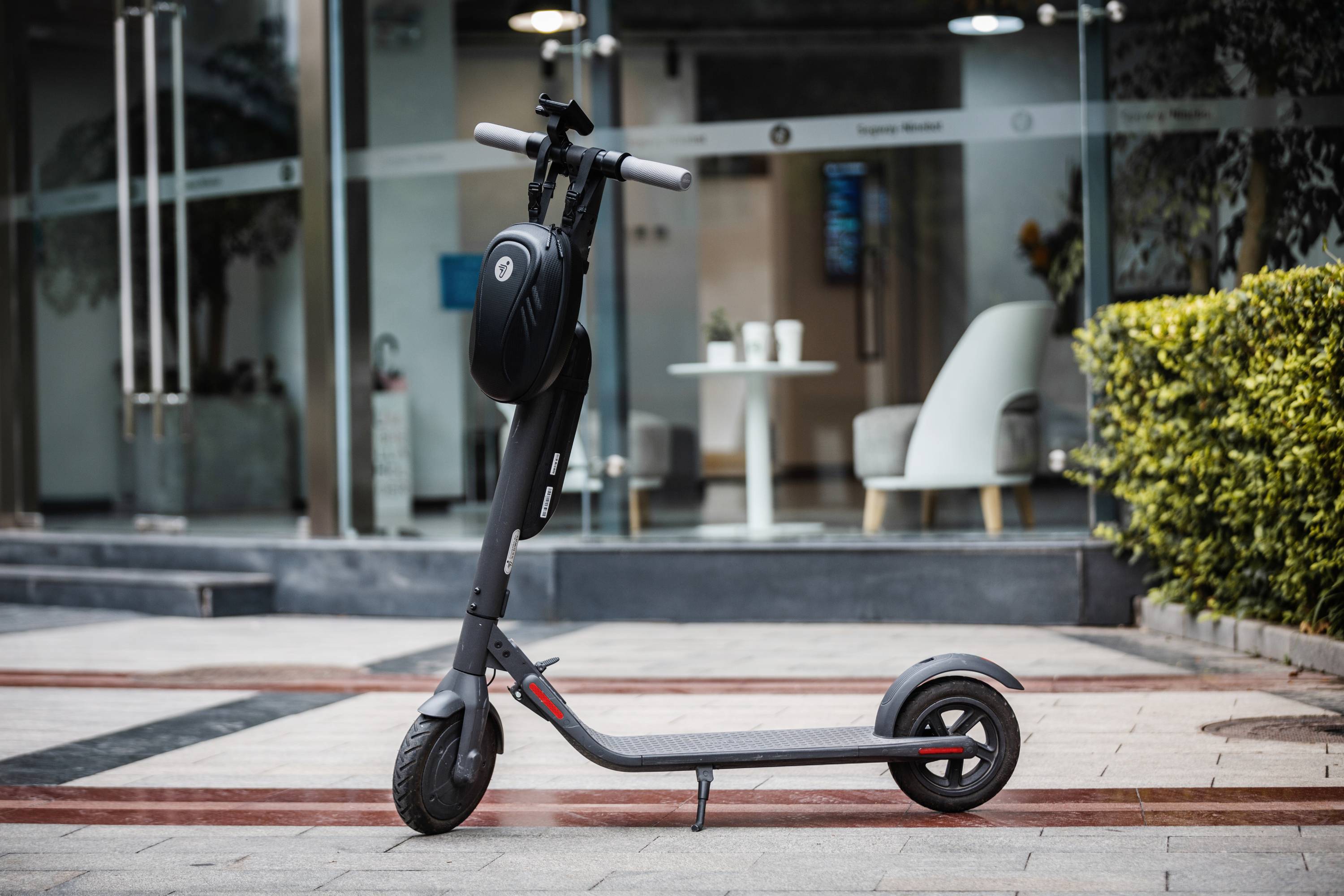 Schwarzer E-Scooter steht auf einem Gehweg vor einem modernen Gebäude mit Glasfassade. Im Hintergrund sind Pflanzen und weiße Stühle zu sehen.