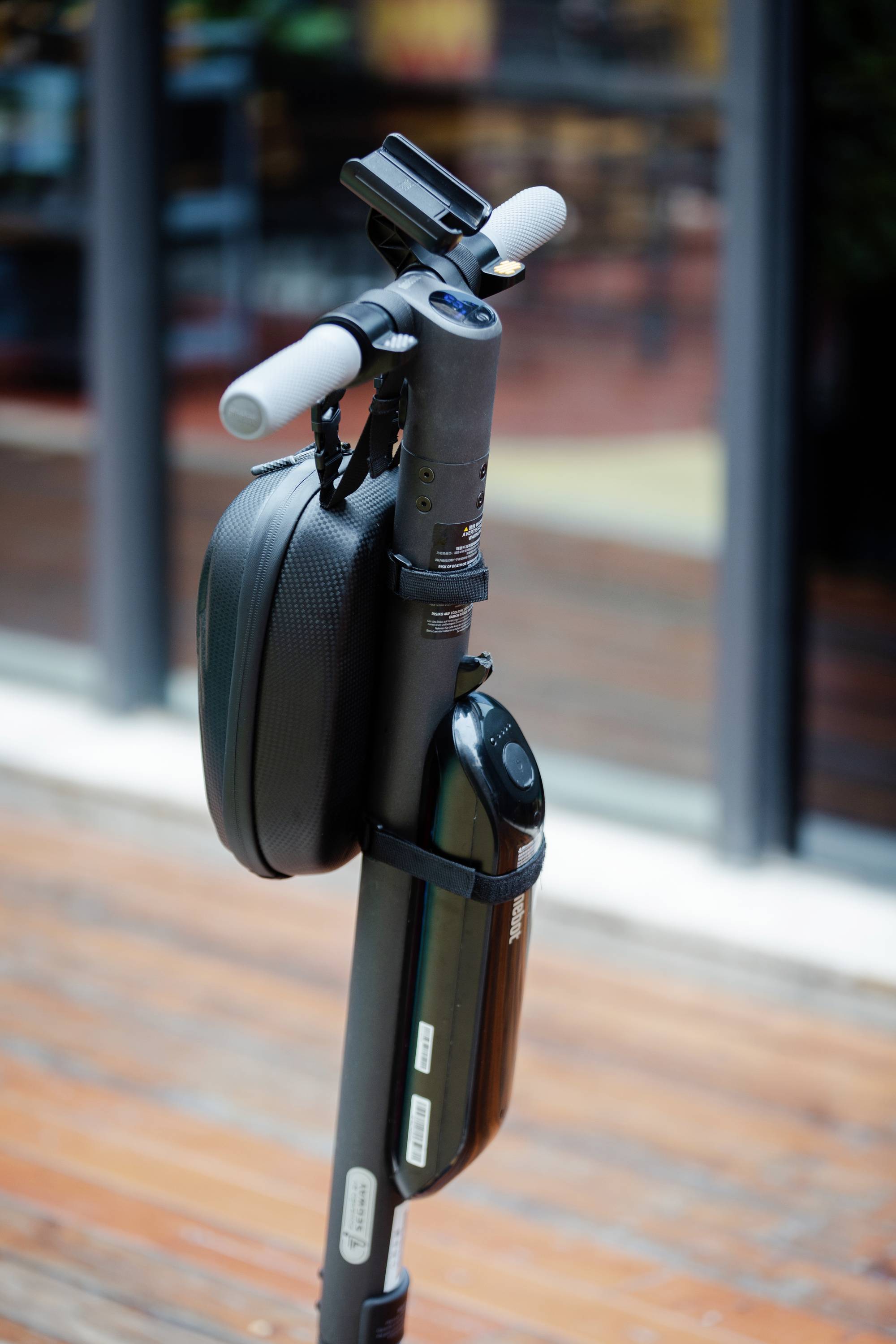 Ein zusammengeklappter elektrischer Roller steht auf einem Gehweg. Er hat eine schwarze Tasche am Lenker befestigt. Im Hintergrund befindet sich eine Glasfassade.