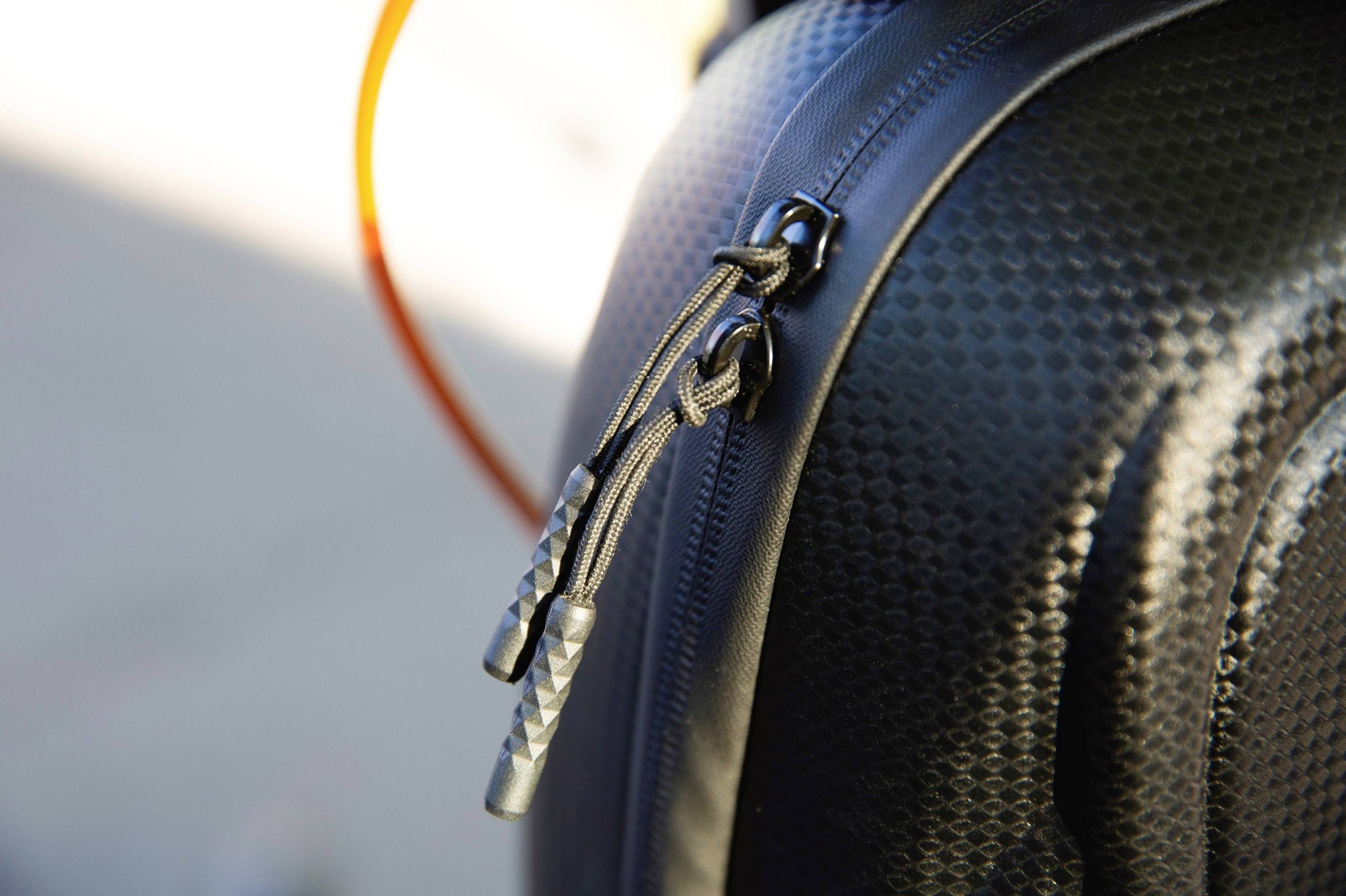 Nahaufnahme eines schwarzen Rucksacks mit Karbonmuster und Reißverschlüssen mit geflochtenen Schlaufen. Hintergrund unscharf.