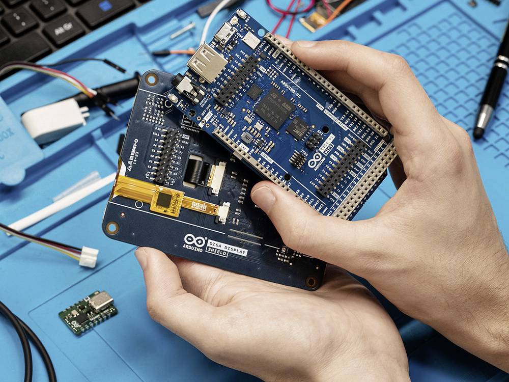 Eine Person hält zwei Mikrocontroller-Boards mit verschiedenen Anschlüssen und Schaltflächen. Im Hintergrund sind Kabel und Werkzeug sichtbar.