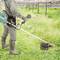 Eine Person in Schutzkleidung benutzt einen Motorsense, um Gras in einem bewachsenen Außenbereich zu schneiden. Im Hintergrund sind Reihen von Weinreben zu sehen.