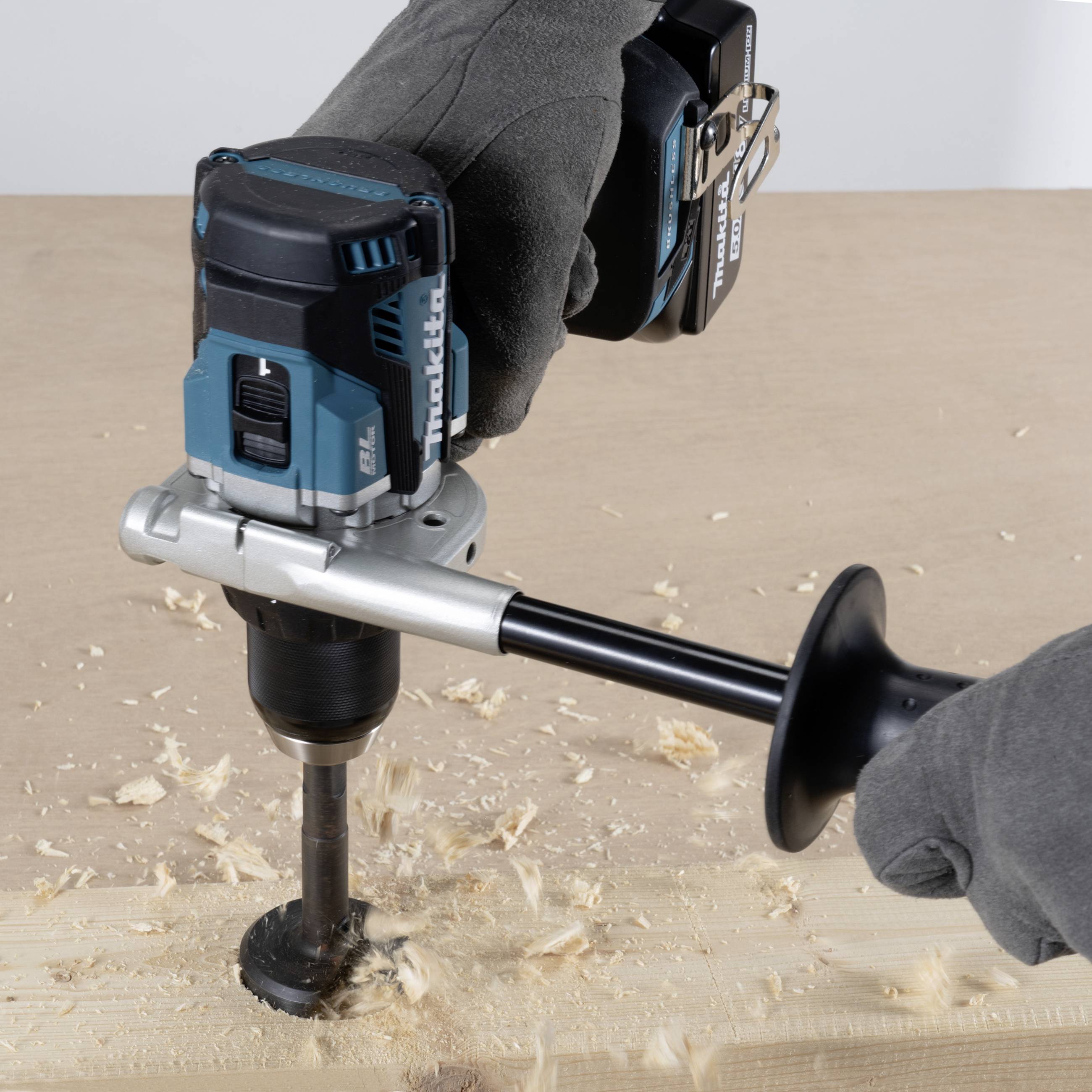 Eine Person in grauen Schutzhandschuhen benutzt eine blaue und schwarze Bohrmaschine, die Holzspäne auf einem Holzbrett erzeugt.