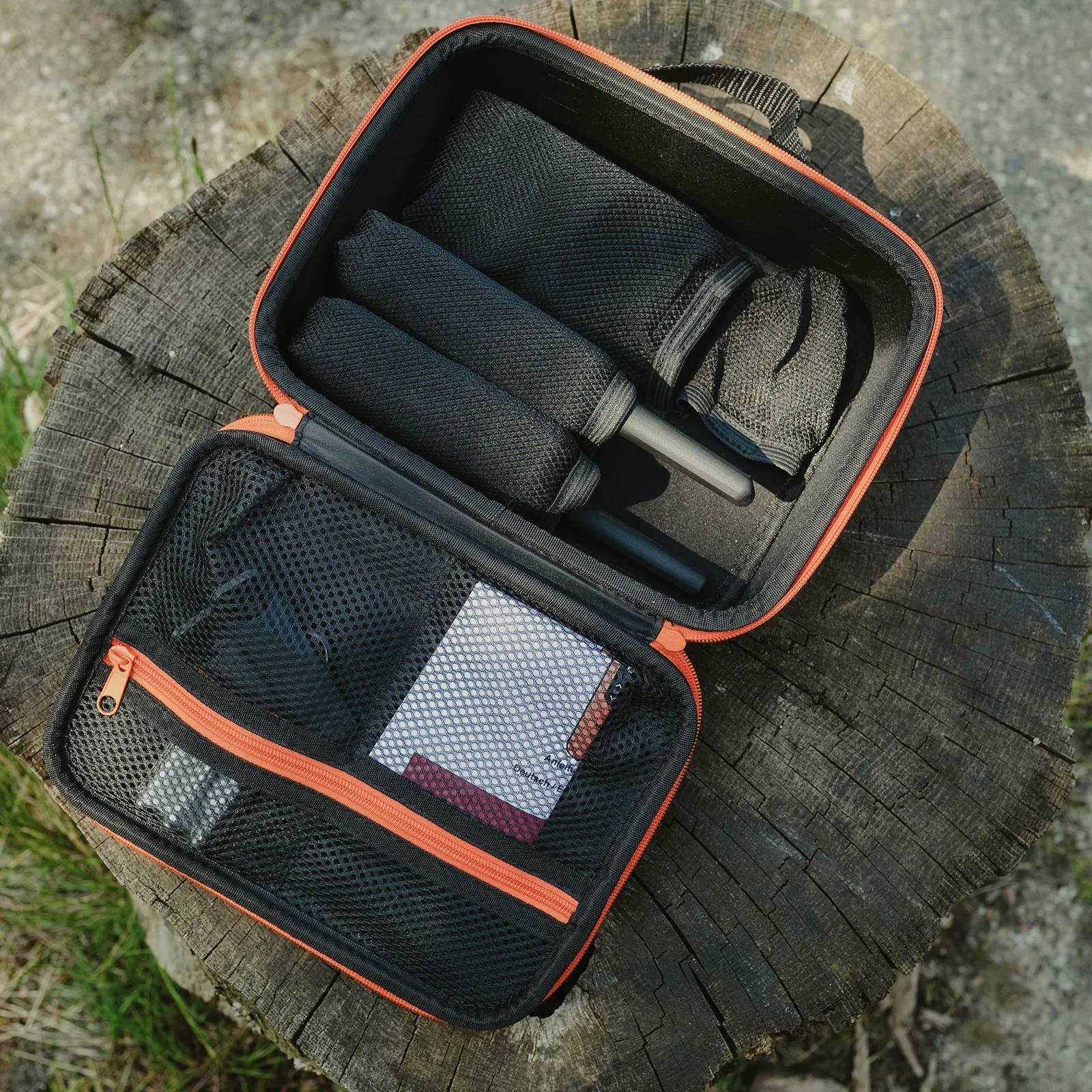 Kompakte schwarze Tasche mit orangefarbenem Reißverschluss. Enthält schwarze Werkzeuge und Netzfächer, liegt auf einem Baumstumpf.