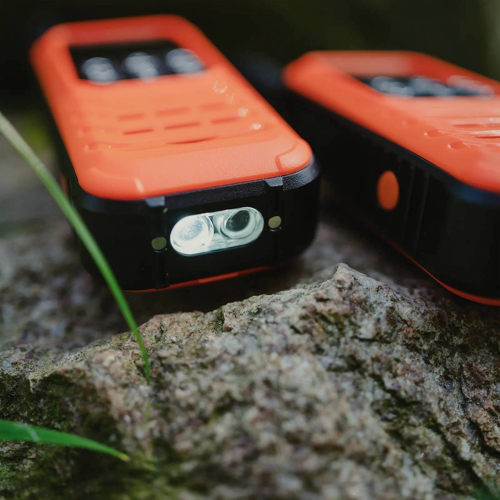 Ein rotes Outdoor-Walkie-Talkie liegt auf einem Felsen. Es wird von einem grünen Grashalm leicht verdeckt.