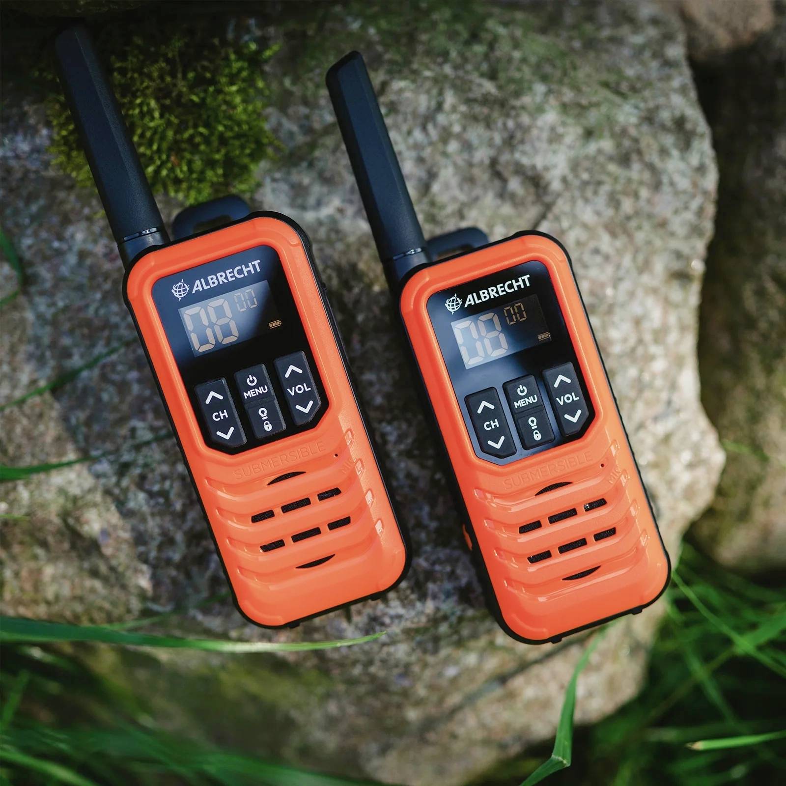 Zwei orangefarbene Walkie-Talkies liegen auf einem Felsen, umgeben von Gras. Sie zeigen unterschiedliche Kanäle auf ihren Displays an.