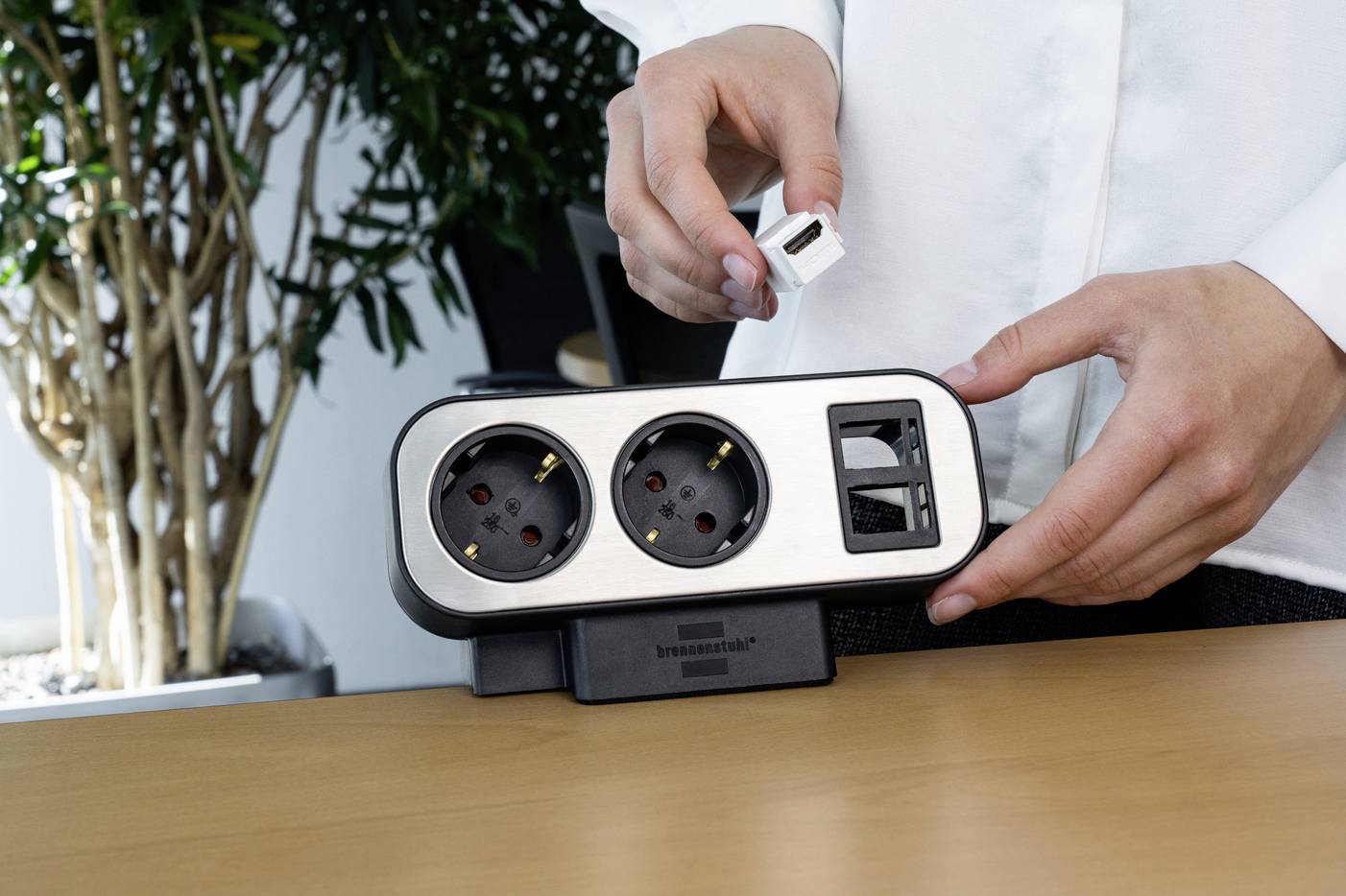 Eine Person zeigt eine Tischsteckdosenleiste mit zwei Steckdosen und zwei USB-Ports. Im Hintergrund ist eine Pflanze sichtbar.