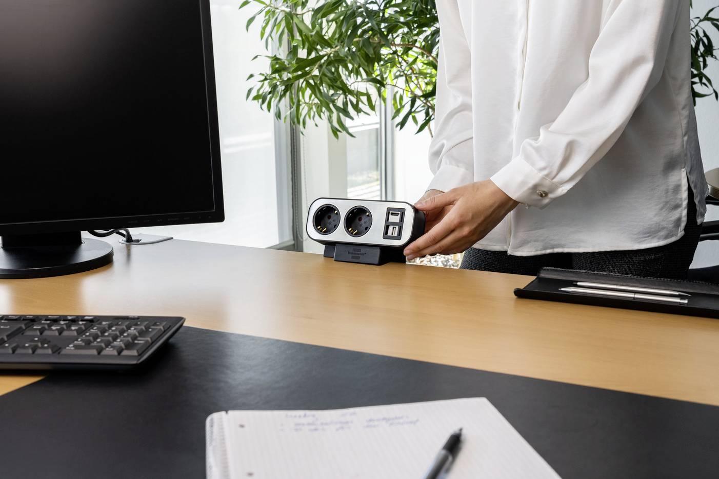Eine Person positioniert eine Steckdosenleiste auf einem Bürotisch. Im Hintergrund sind Pflanzen und ein Computerbildschirm sichtbar.