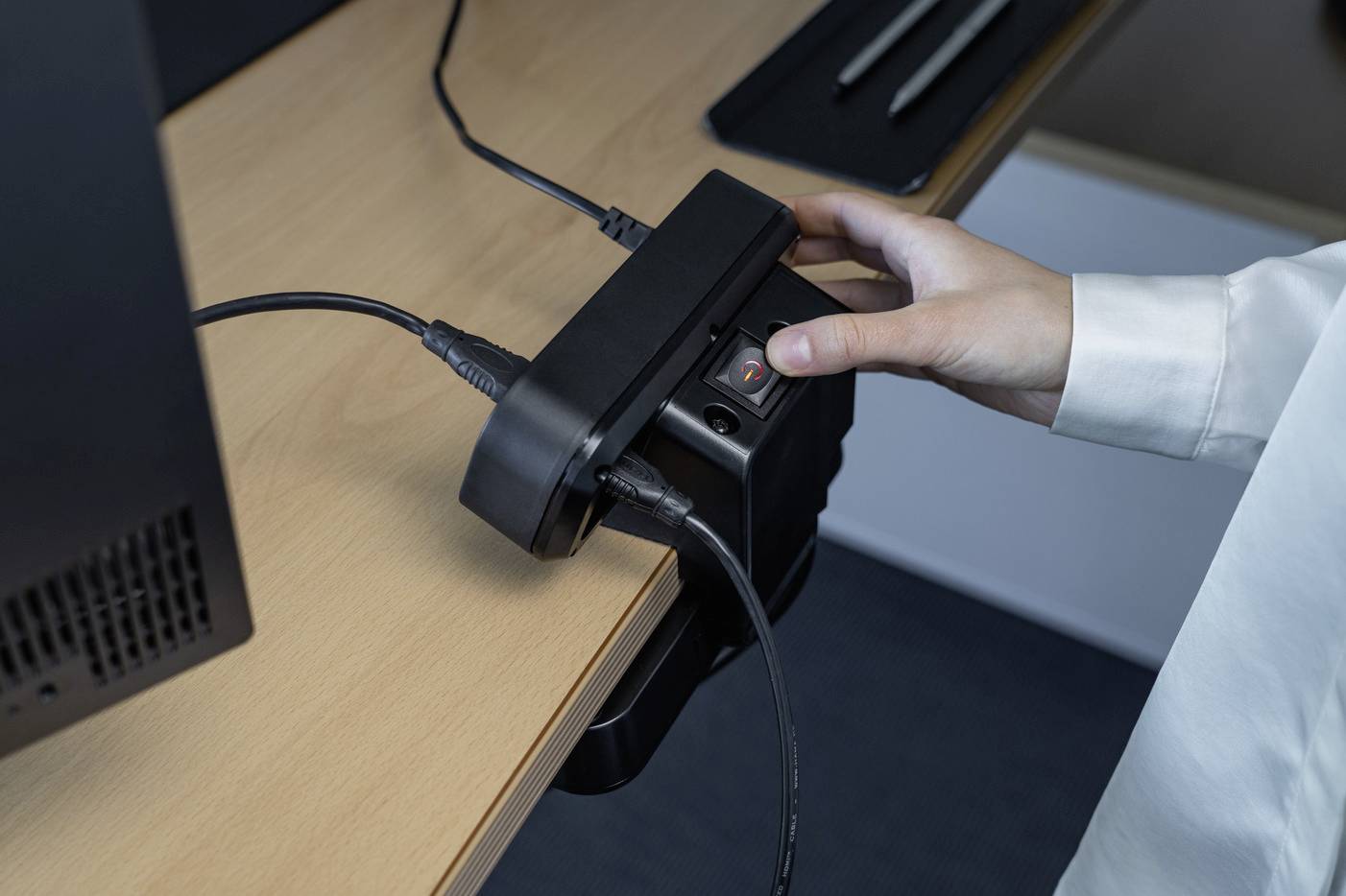 Eine Hand bedient ein elektronisches Gerät an einem Schreibtisch, das mehrere Kabel angeschlossen hat, möglicherweise zur Stromversorgung.