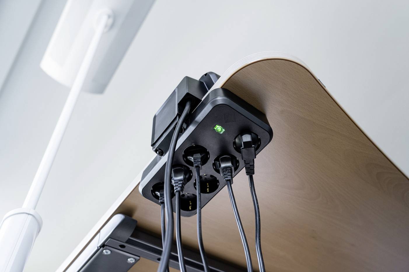 Mehrere Kabel sind an einen Mehrfachstecker unter einem Tisch angeschlossen. Ein grünes Licht zeigt an, dass die Steckerleiste aktiv ist.